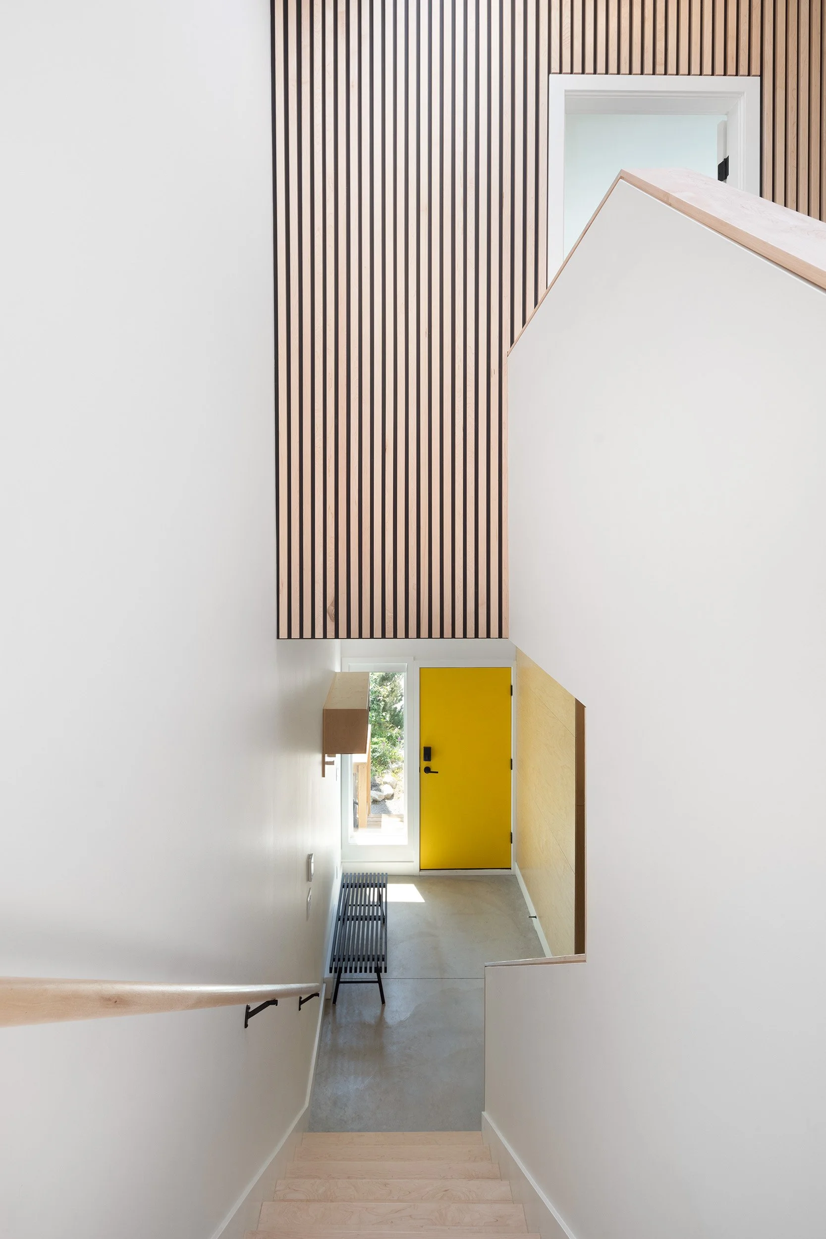 Interior view of a modern building with a staircase, white walls, a yellow door, and wooden accents on the wall and staircase railing.