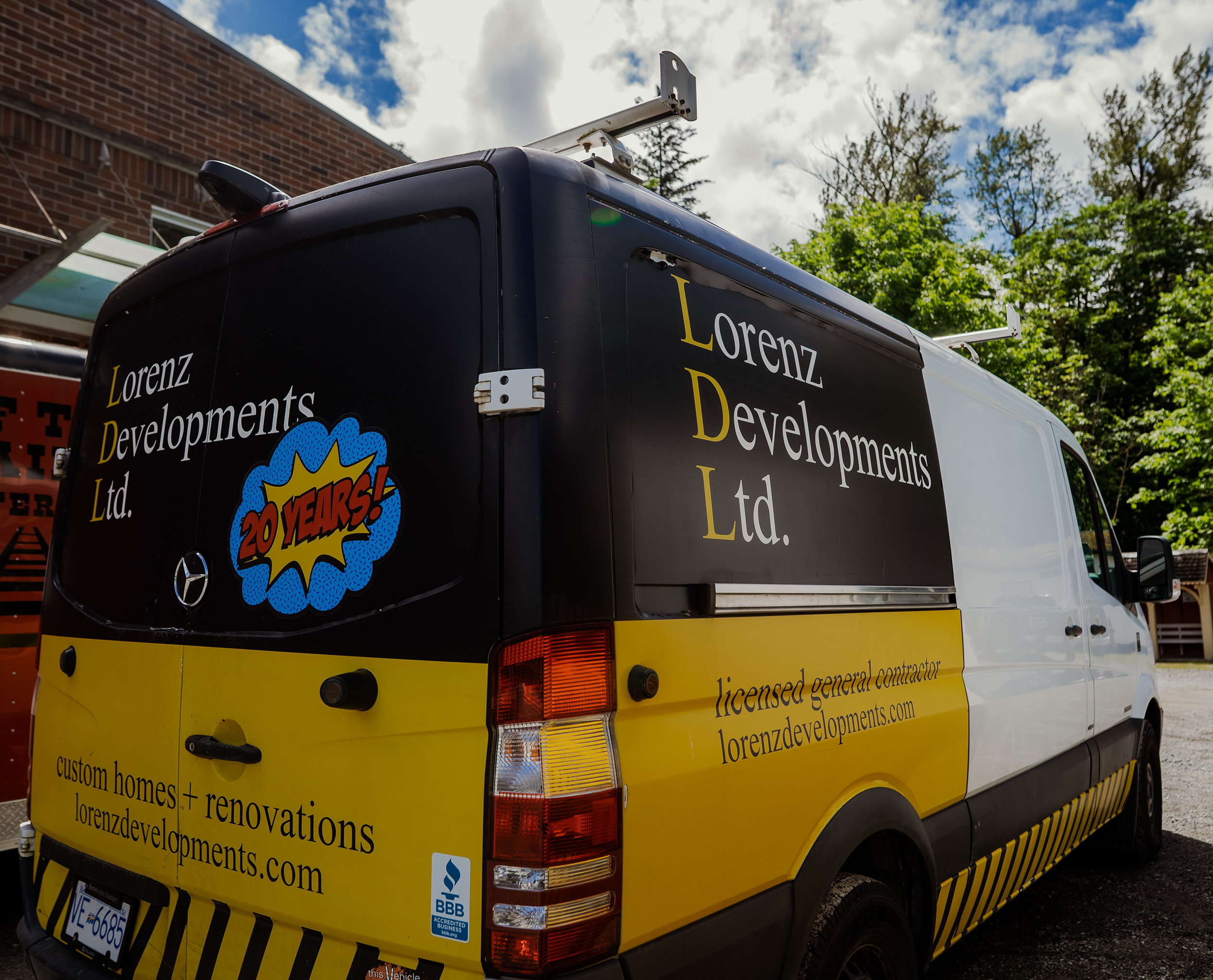 A delivery van with black and yellow coloring and the logo "Lorenz Developments Ltd." on the side. The van celebrates 20 years with a colorful comic-style burst graphic. It is parked outdoors, against a backdrop of green trees and a partly cloudy sky.