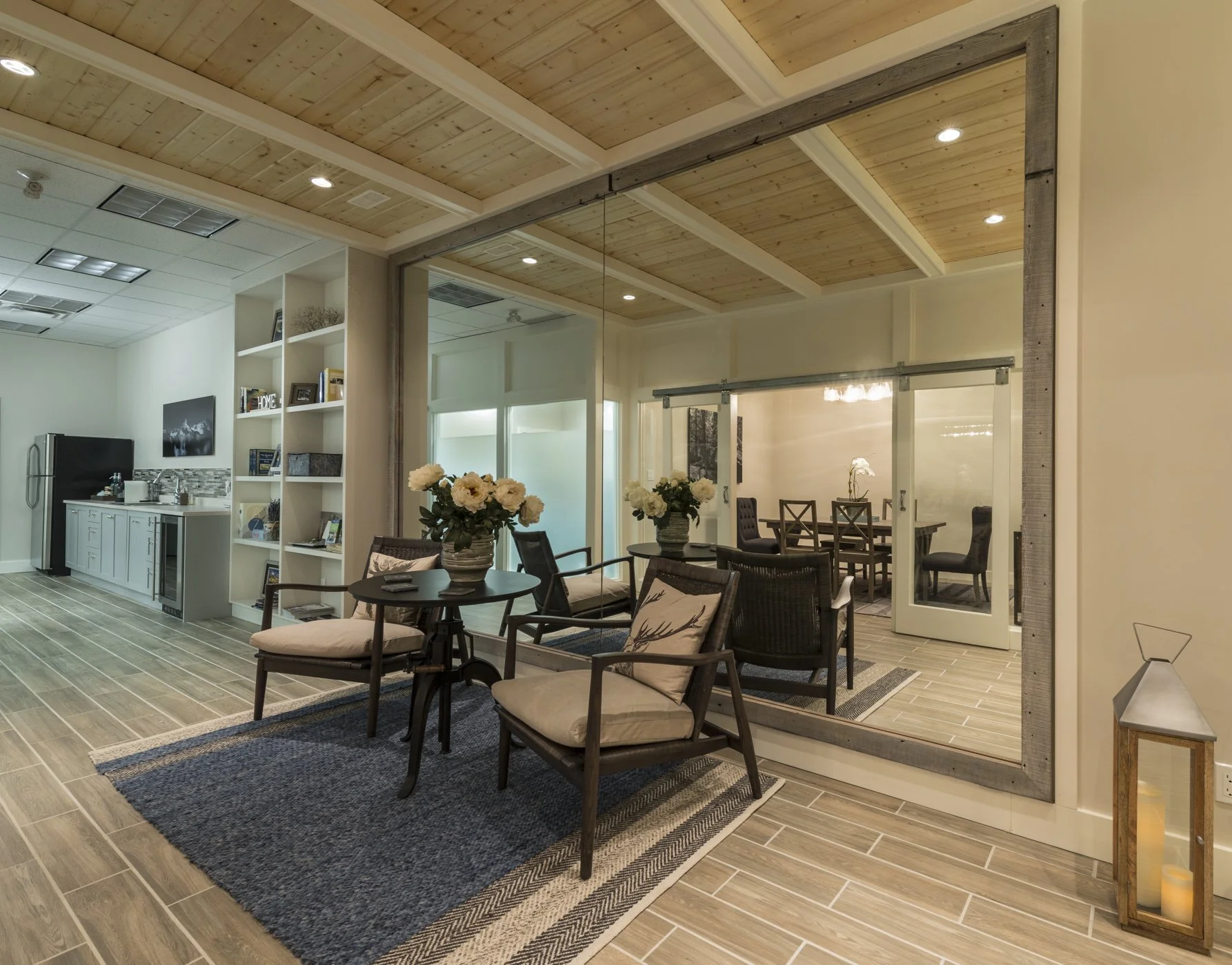 Indoor lounge area with chairs, coffee table, flower vases, and a sliding glass door leading to a dining room with a wooden table and chairs. The ceiling is wooden with recessed lighting, and the room has wood-look tile flooring.