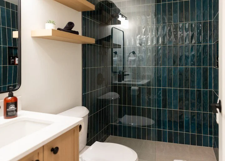 Bathroom with dark blue tiled shower area, wooden shelves, white toilet, white sink, and mirror.