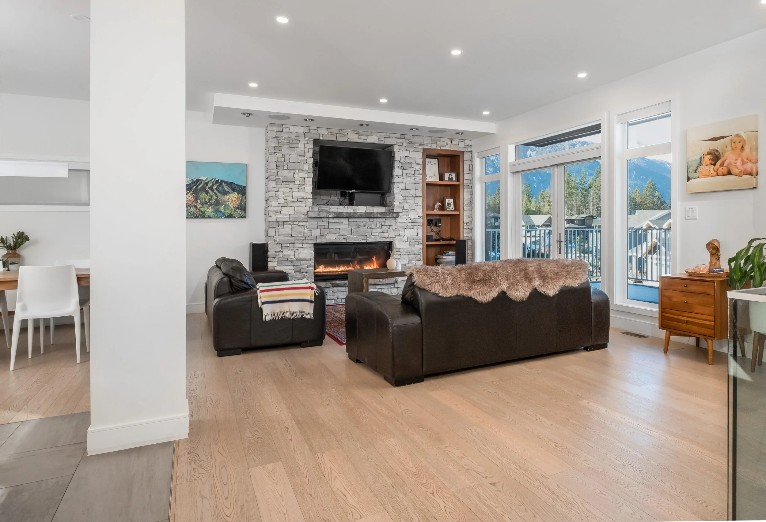 A modern living room with a stone fireplace, a flat-screen TV mounted above it, and large windows showing a mountain landscape outside. There are two black leather sofas with a furry throw, a wooden side table, and various art pieces on the walls.