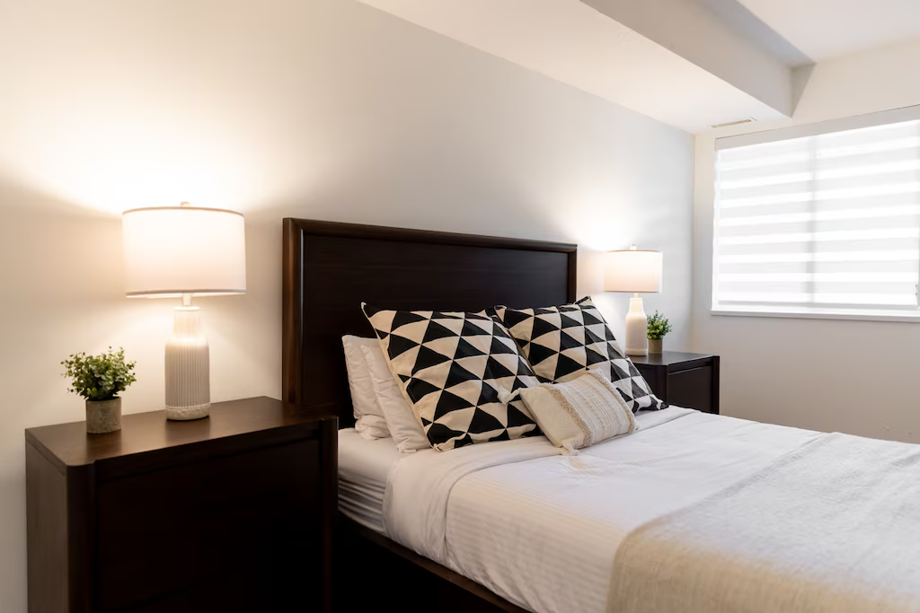 A neatly made bed with a dark wooden headboard, patterned black and white pillows, and a small decorative pillow, flanked by matching nightstands with table lamps and potted plants, in a minimalist bedroom with a window with horizontal blinds.