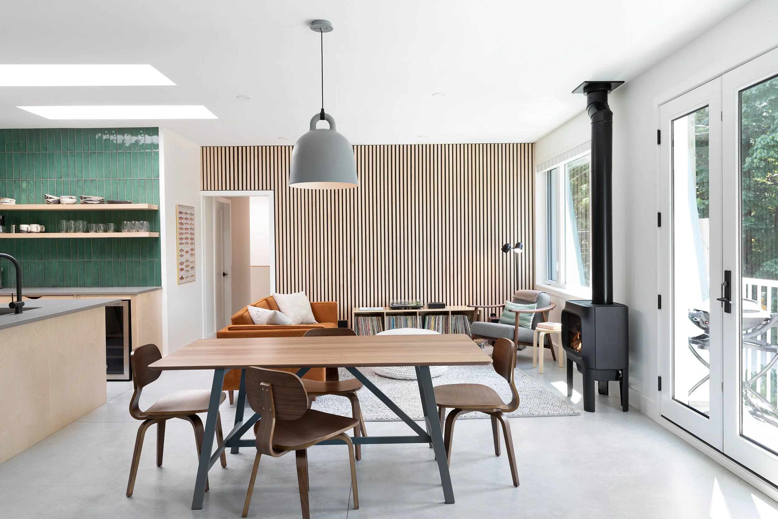 A modern open-concept living room and kitchen with large windows, a wood dining table with chairs, a fireplace, green tiled wall, and a mix of wooden, gray, and orange furniture.