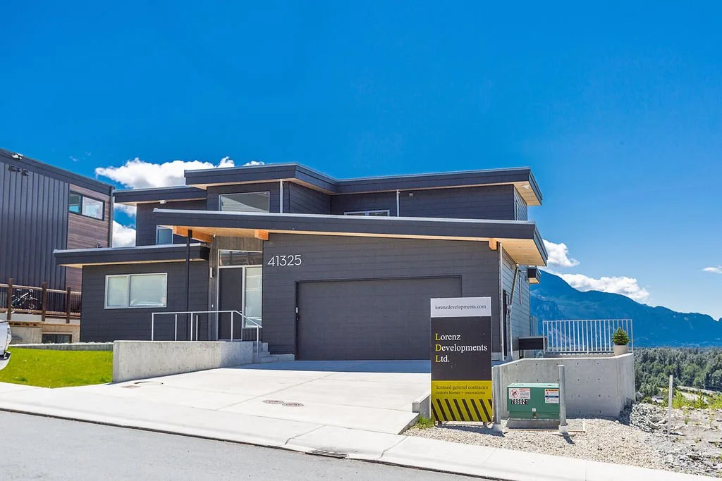 New modern house with dark gray exterior, situated in a mountainous area, under a bright blue sky with some clouds.