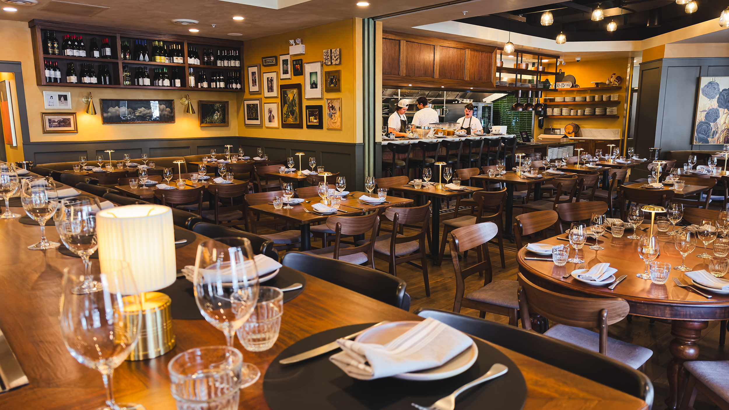 Inside a modern restaurant with tables set for dining, including glasses, napkins, and cutlery. The background features a bar area with wine bottles and a kitchen with chefs preparing food. The decor includes framed art on yellow walls and warm light