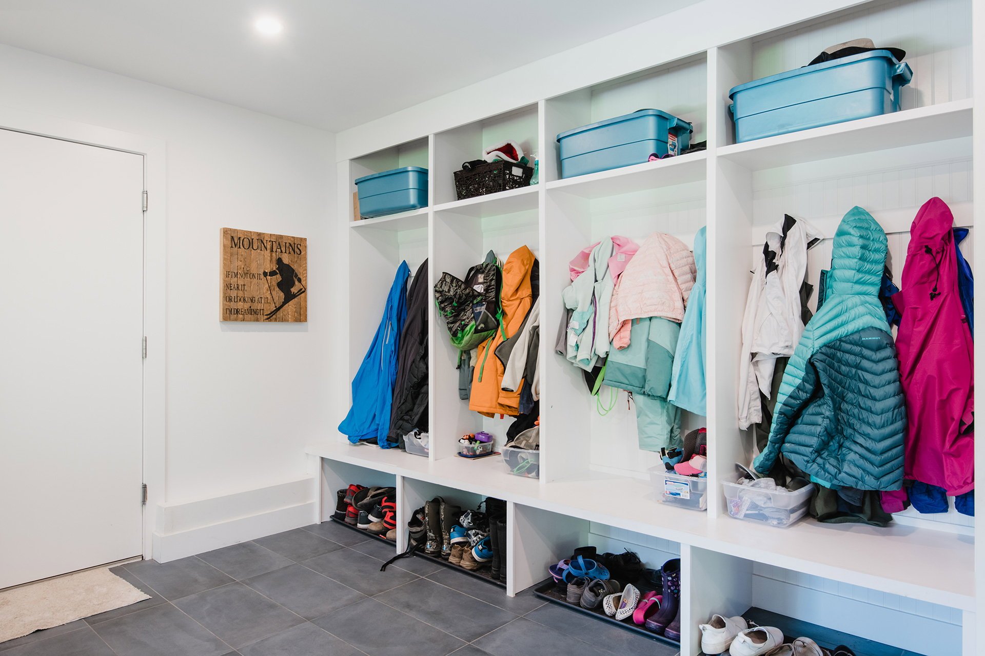 A closet with white shelves containing jackets, shoes, and storage containers, with a sign on the wall reading 'Mountains.'