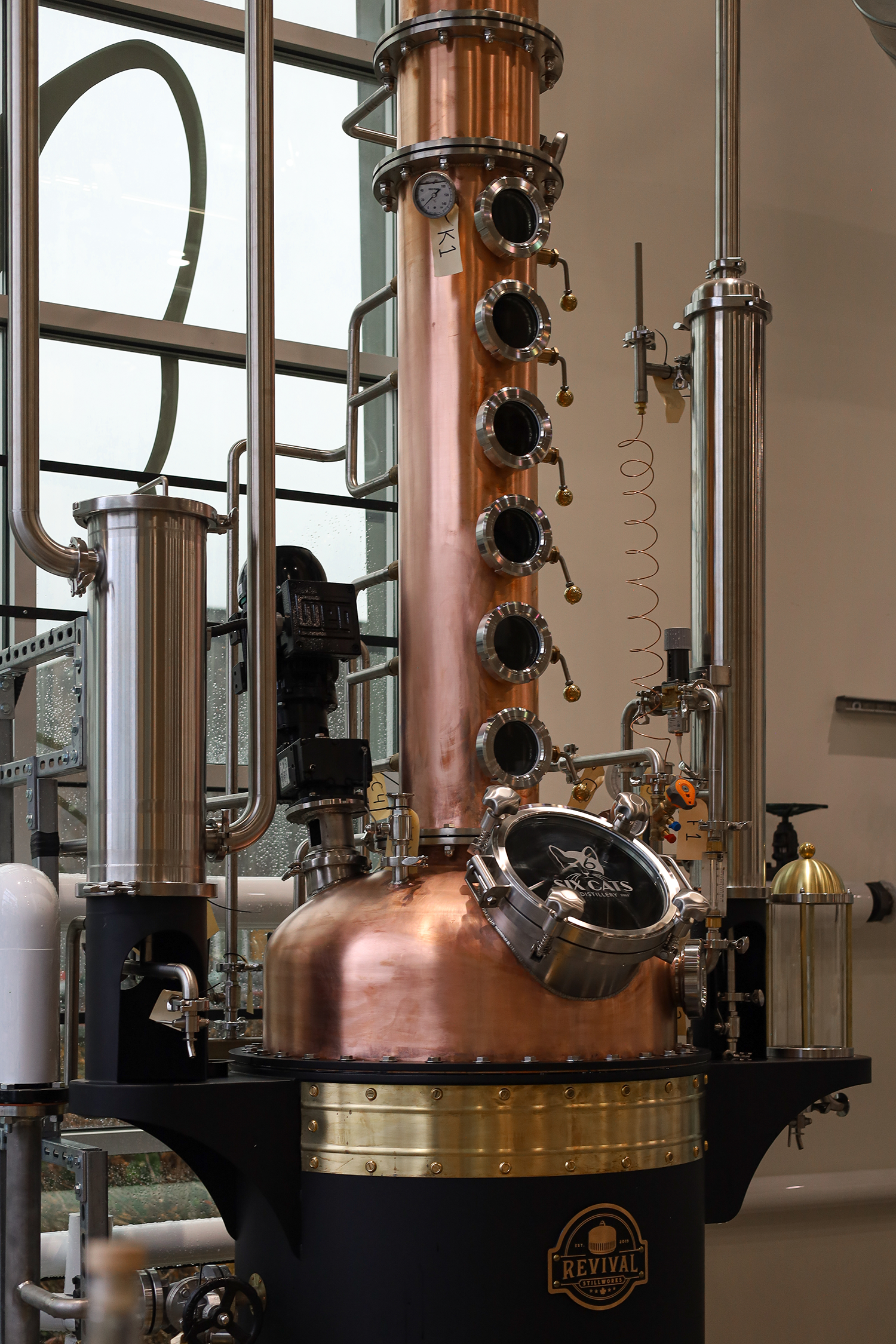 A large copper distillation still with multiple sight glasses, gauges, and pipes, situated inside a modern building near large windows.