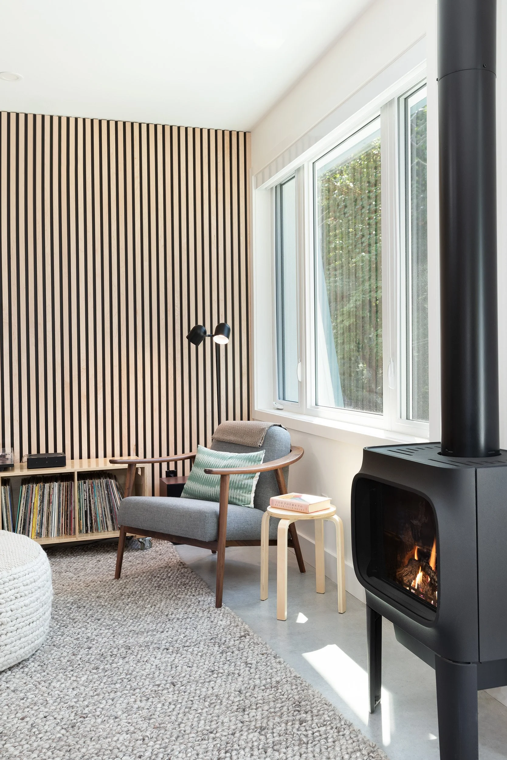 A cozy living room corner with a wood-paneled accent wall, a gray armchair with a stripe pillow, a small side table with books, a fireplace with a black stove pipe, and large windows overlooking a green outdoor area. A woven rug covers the floor.