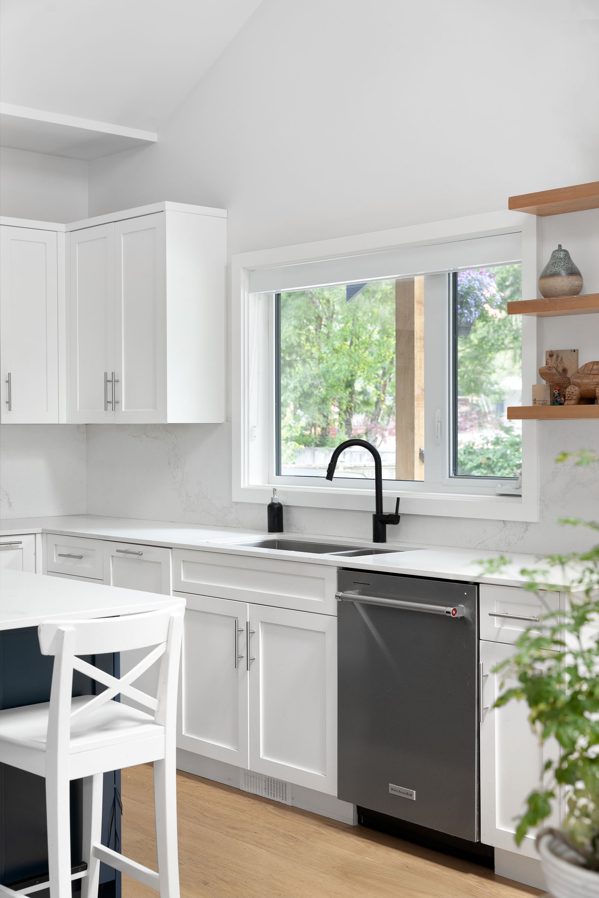 Modern kitchen with white cabinets, black faucet, window overlooking green trees, and wooden shelves with decorative items