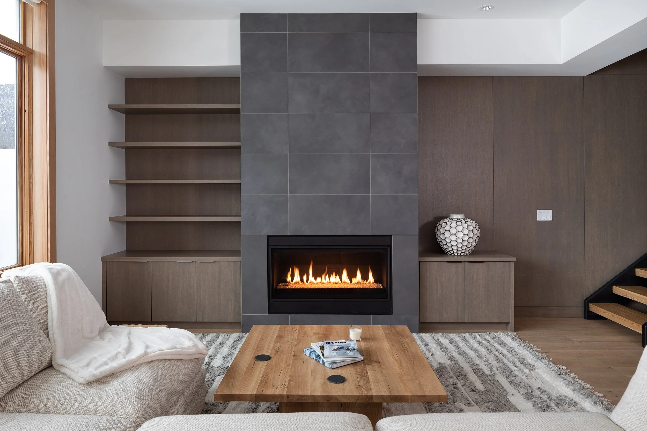 Modern living room with a fireplace, beige sofa with a blanket, wooden coffee table with magazines, gray tiled wall, wooden shelves, decorative vase, and staircase to the right.