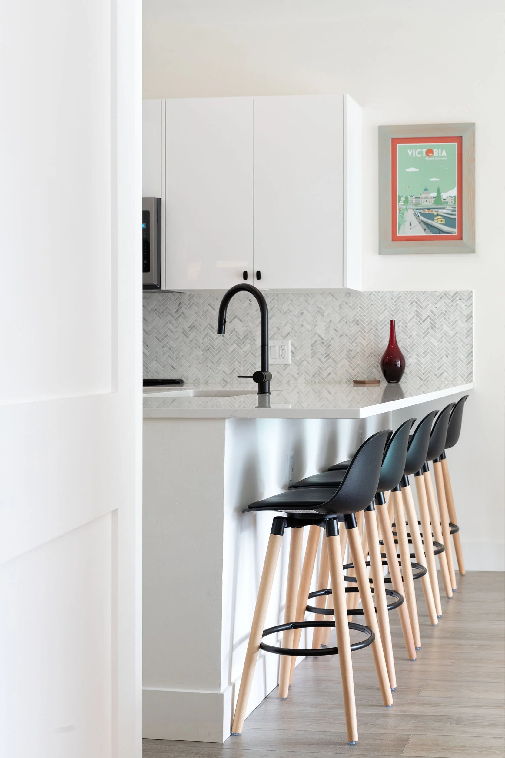 Modern kitchen with a white counter, black bar stools with wooden legs, a black faucet, white upper cabinets, a red vase, and a framed poster of Victoria, Australia, on the wall.