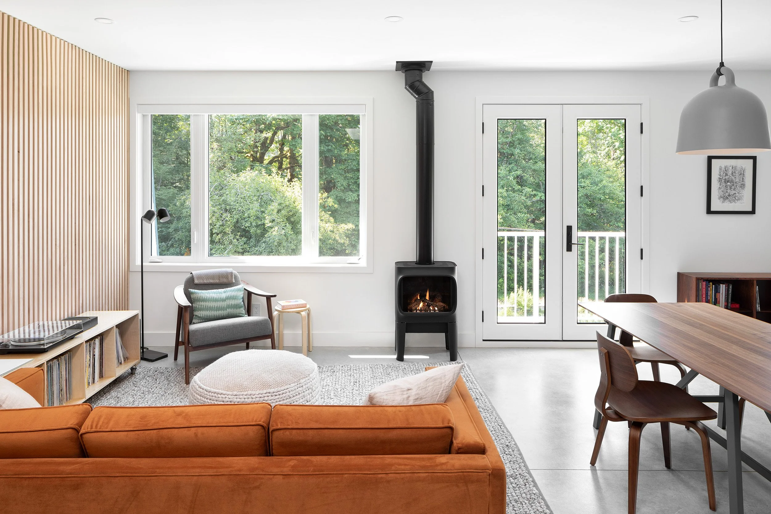 A living room interior with a large orange velvet sofa, a gray armchair with a patterned cushion, a wooden side table, a black wood stove with a chimney pipe, large windows showing green trees, and a wooden dining table with chairs. There's also a fr