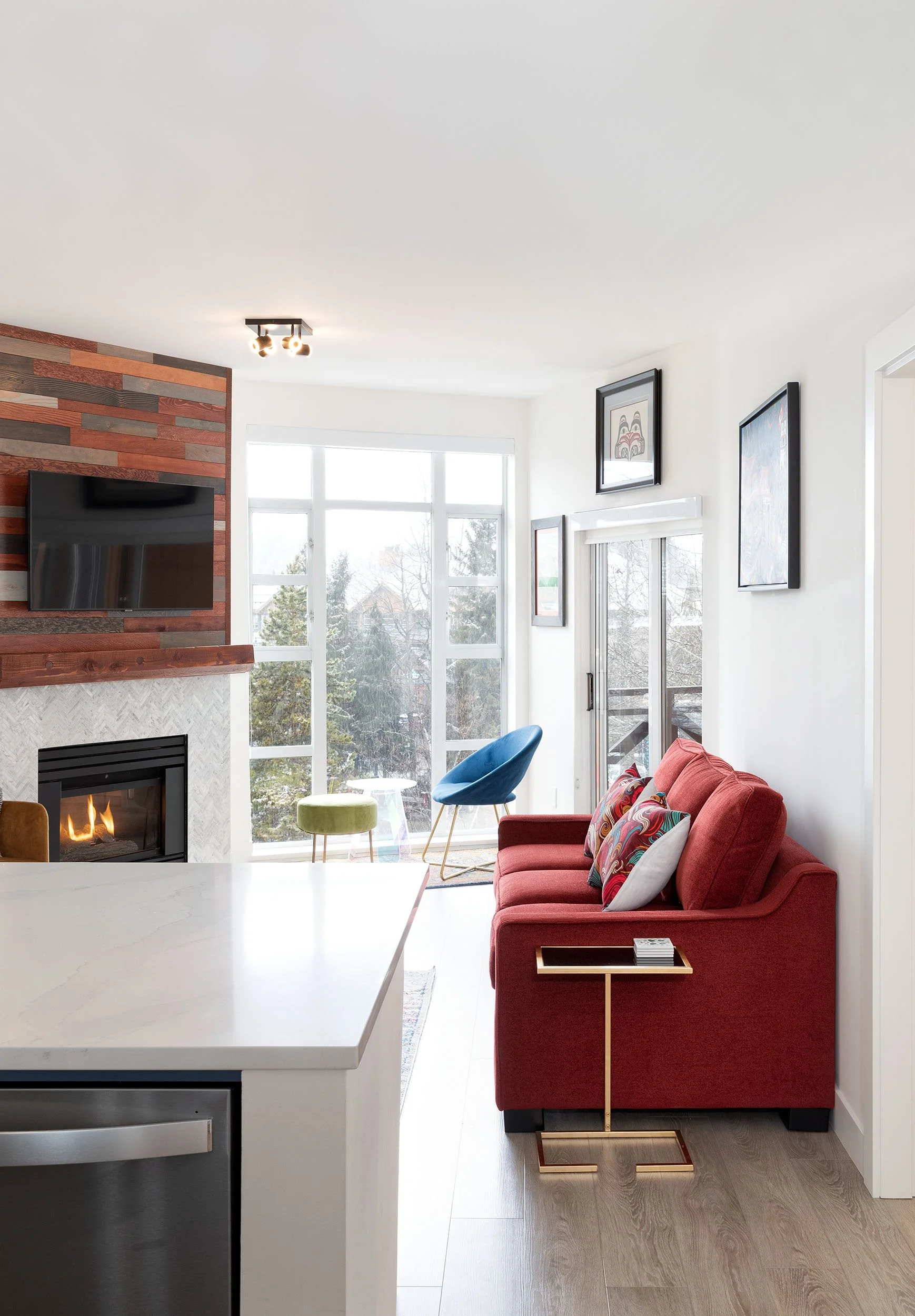 A modern living room with large windows, a red sofa with colorful pillows, a blue armchair, and artwork on the white walls. A fireplace is visible on the left, and natural light fills the space.