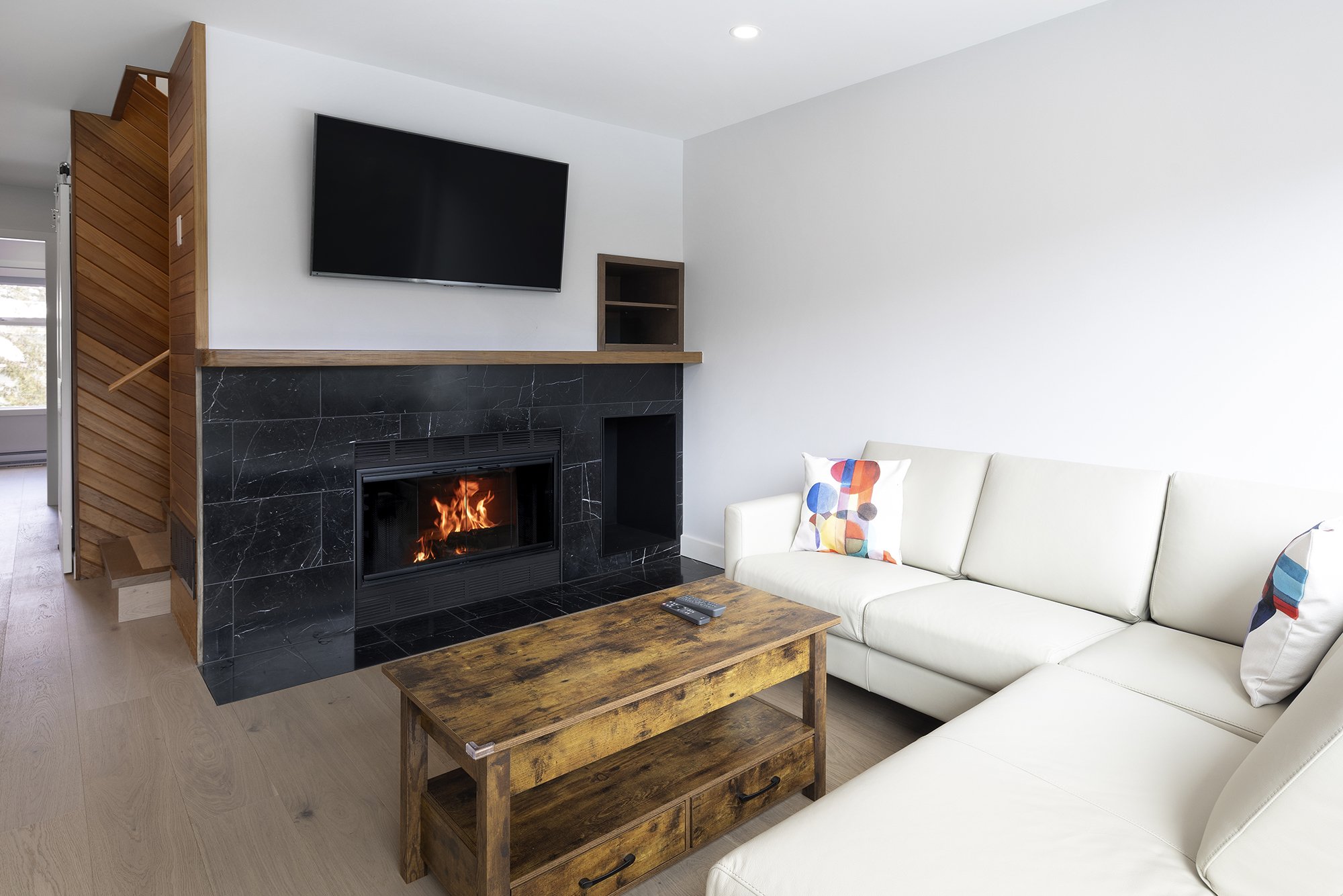 Living room with a fireplace, television, white sofa with colorful patterned pillows, wooden coffee table, and a black TV