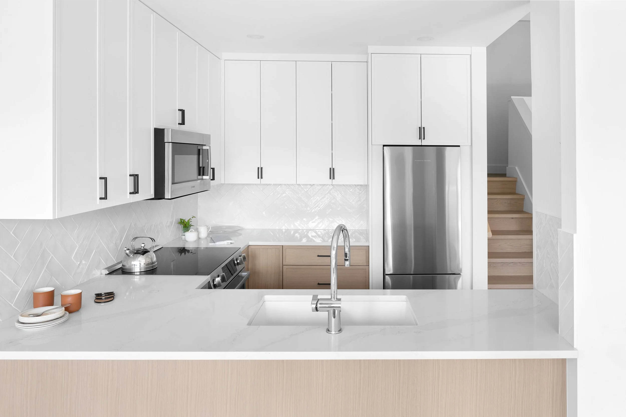 Modern kitchen with white cabinets, stainless steel microwave and refrigerator, white marble countertop, and wooden stairway in the background.