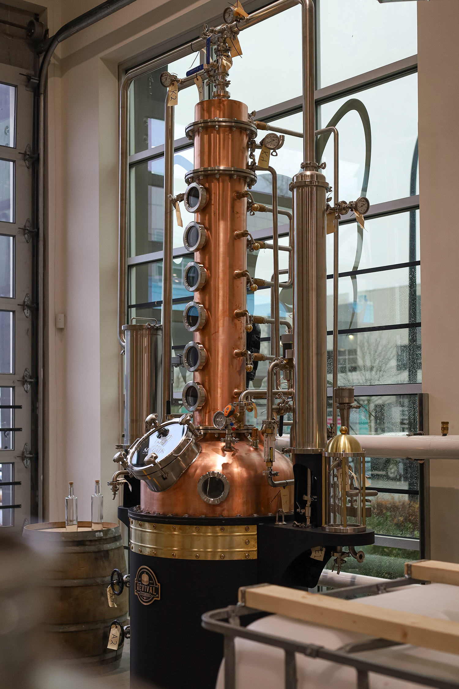 A large copper distillation apparatus inside a building with large glass windows, surrounded by bottles and wooden barrels.