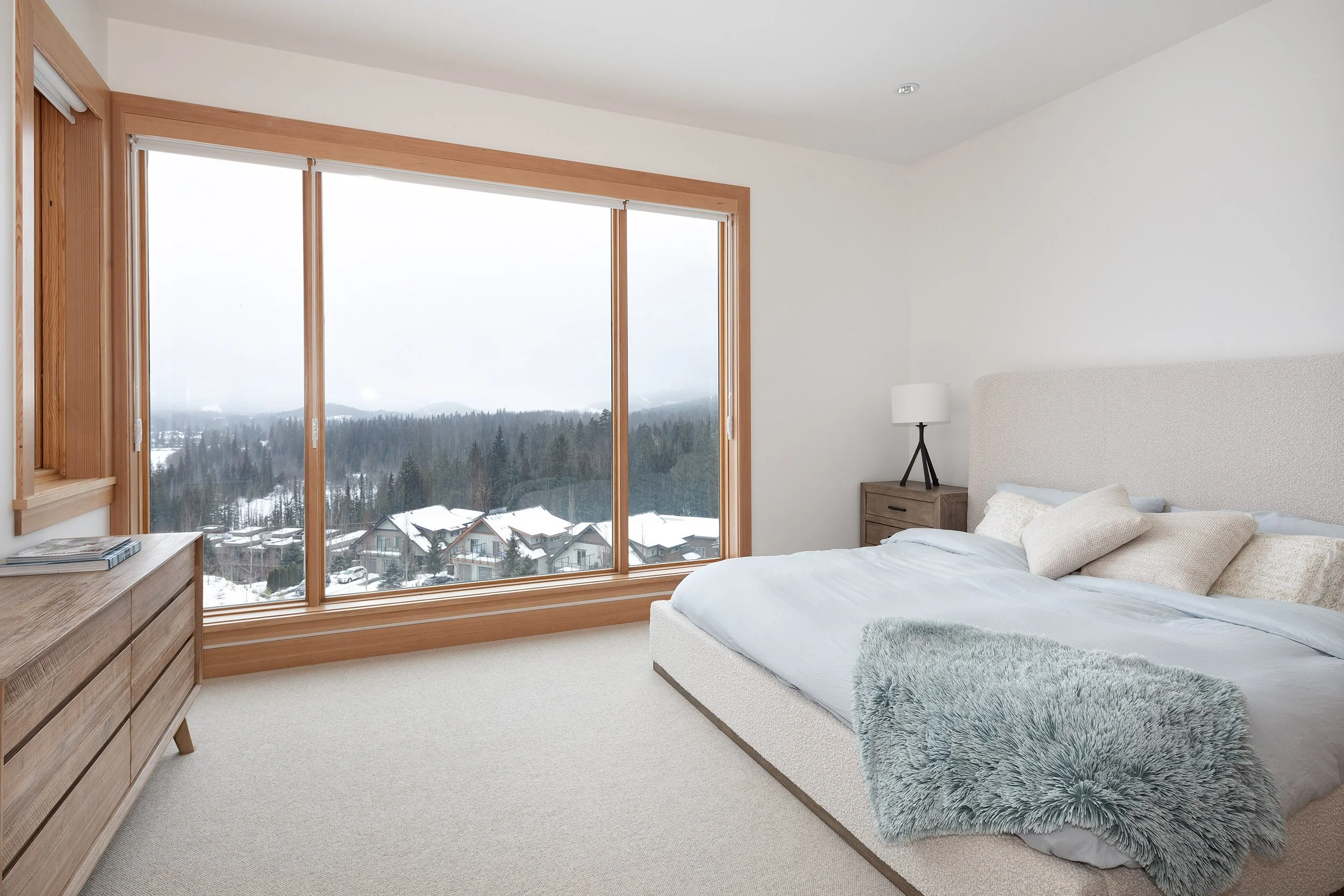A bedroom with a large window showing snow-covered houses and a forested mountain landscape outside. The room has a white bed with beige pillows, a wooden nightstand, and a modern lamp.