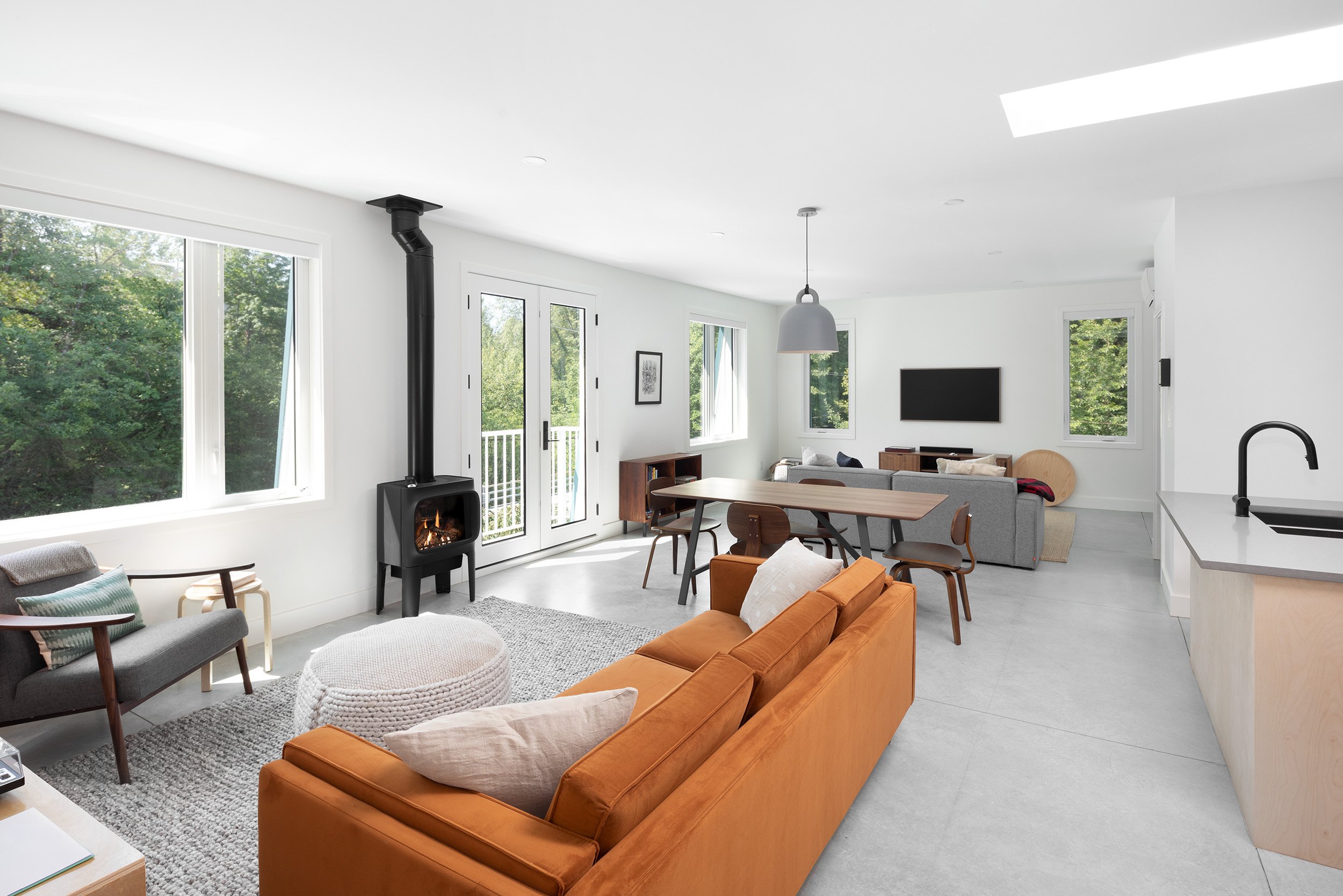 Modern living room with large windows, white walls, a wood stove, a gray sofa, a wooden dining table with chairs, and a kitchen area with a black faucet.