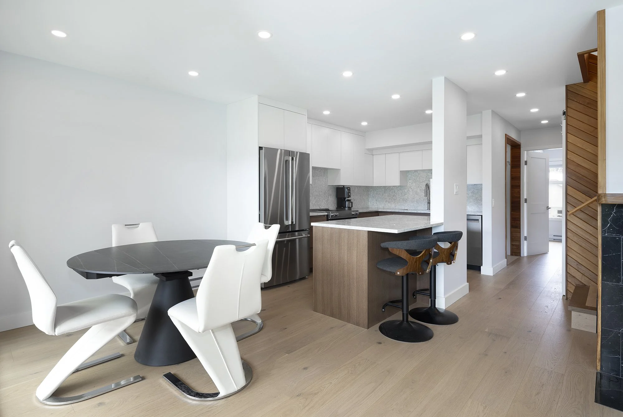 Modern kitchen and dining area with white walls, wooden floors, a black round dining table with white chairs, a kitchen with white cabinets, stainless steel refrigerator, gray countertop and backsplash, kitchen island with bar stools, and wooden acce