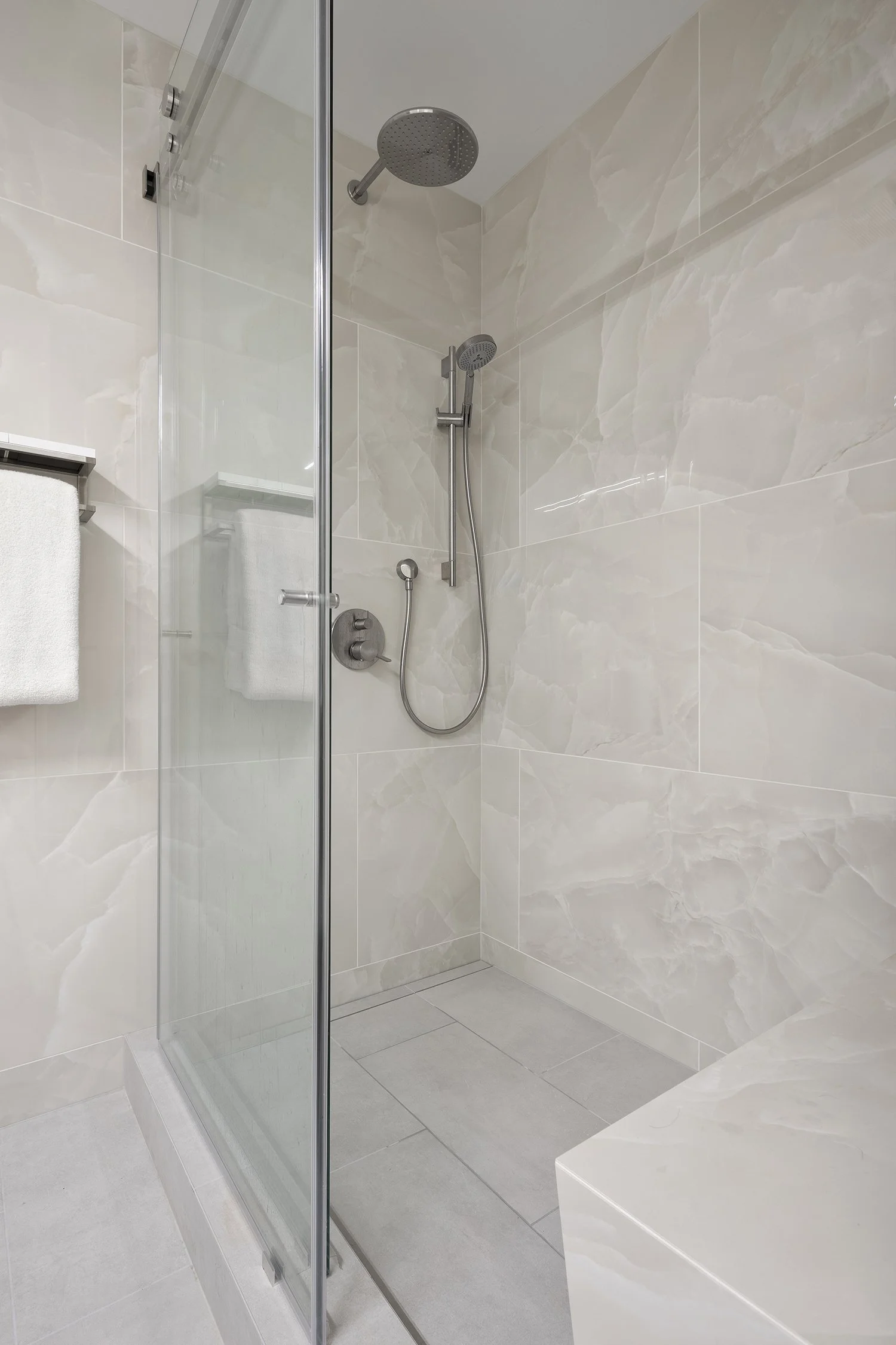 A modern shower area with beige marble tiles, a glass enclosure, and reaching two showerheads—one fixed and one handheld—mounted on the wall.