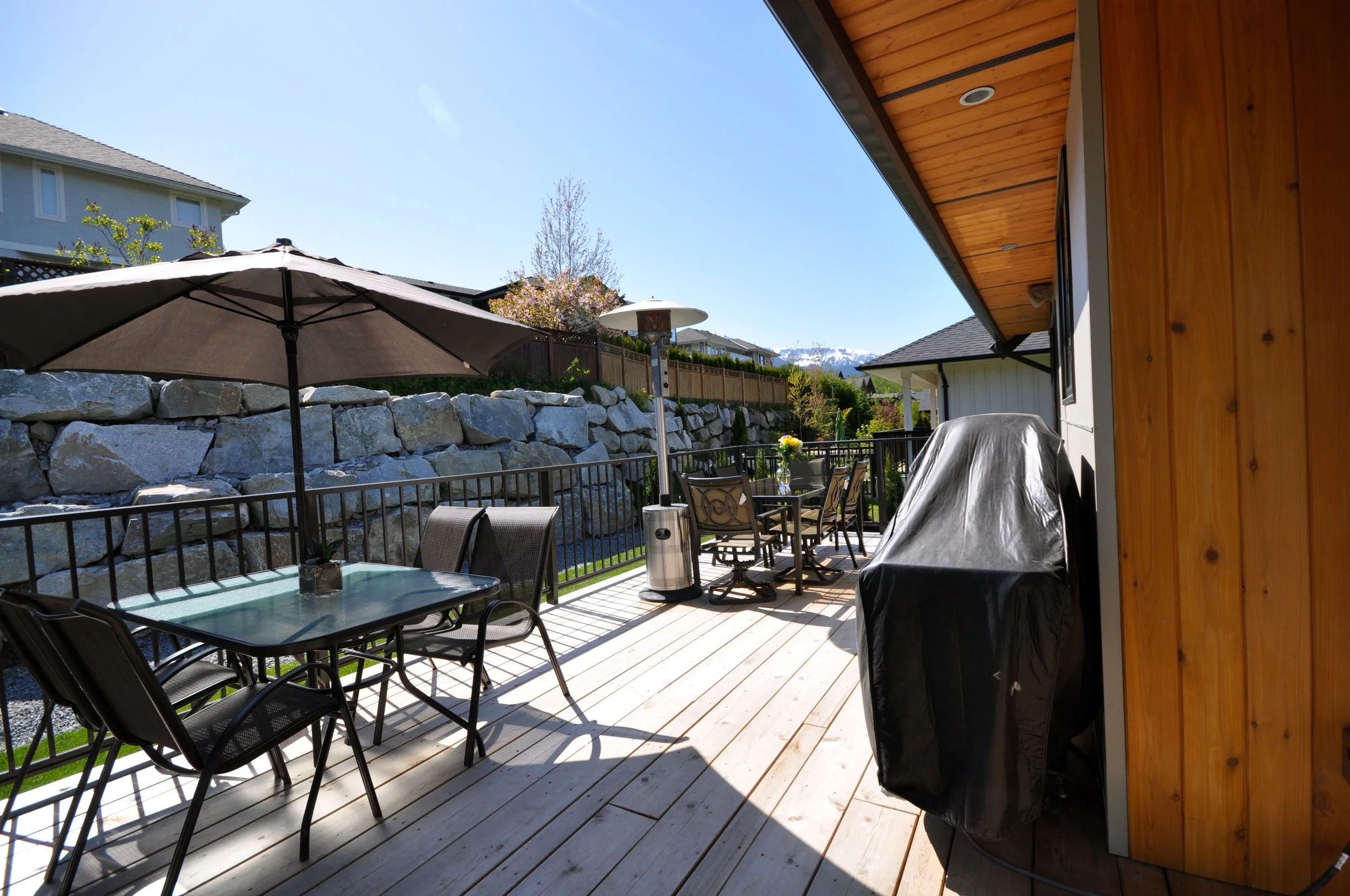 A backyard patio with outdoor furniture, umbrella, and a gas grill, surrounded by a stone wall and neighboring houses with mountains in the background.
