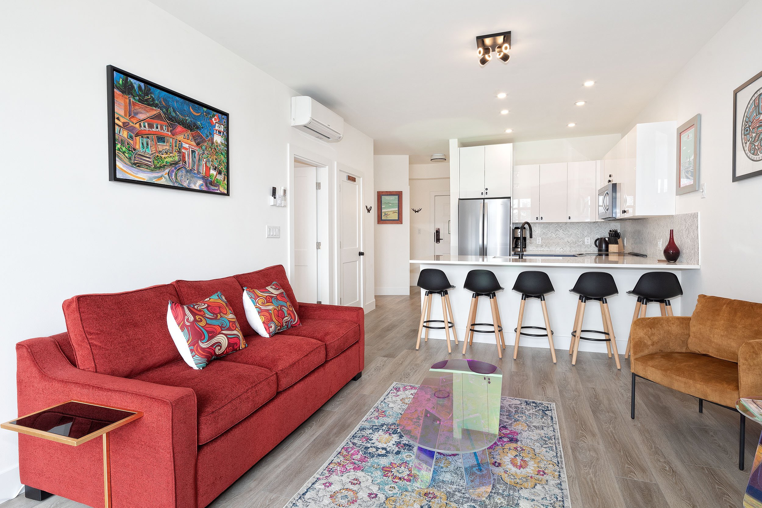 Living room with red sofa, colorful patterned pillows, glass coffee table, and a multicolored rug. Kitchen in the background with white cabinets, stainless steel refrigerator, and a breakfast bar with black stools. Artworks on white walls and wooden 