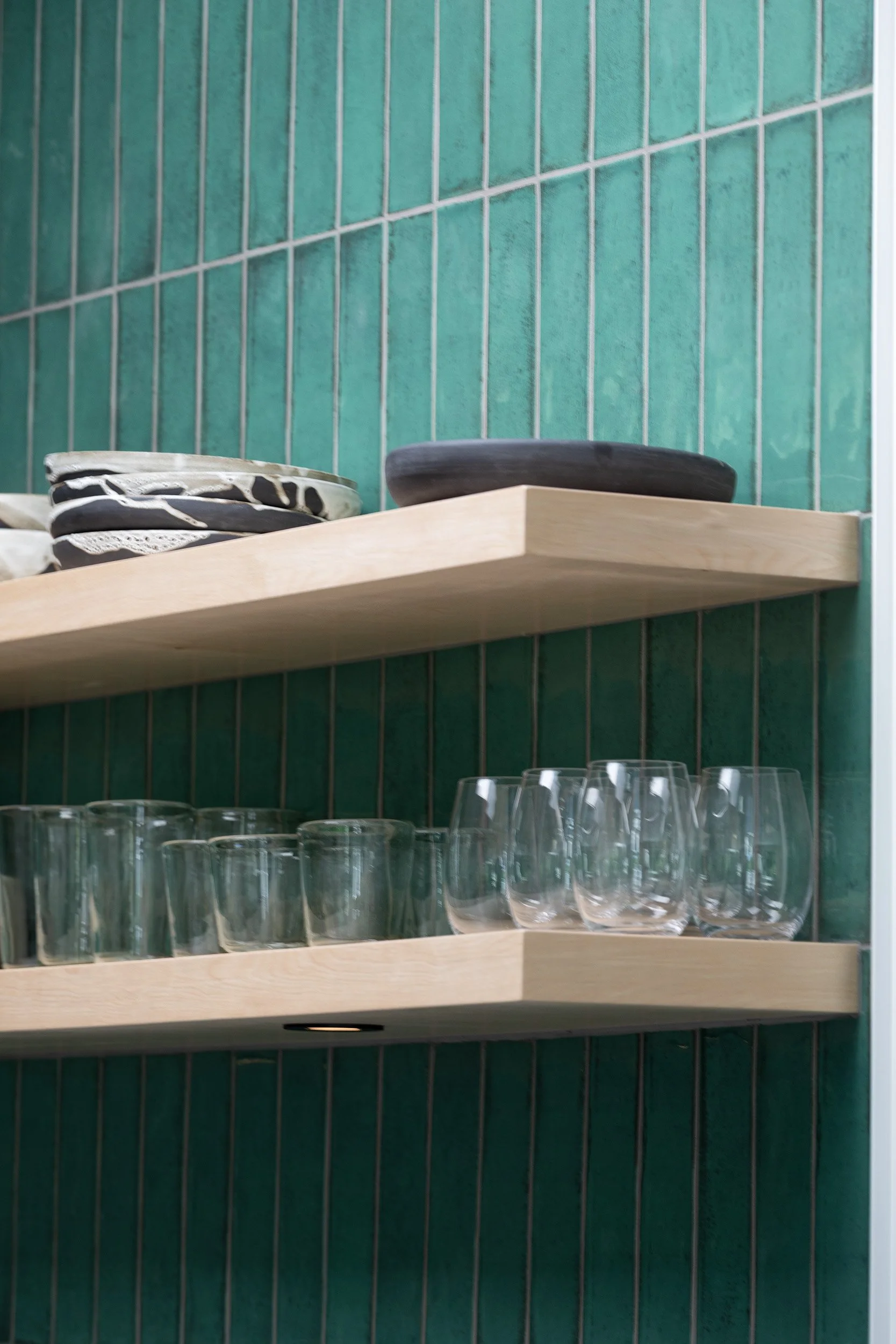 Wooden shelves mounted on a green tiled wall, holding decorative plates and empty glasses.