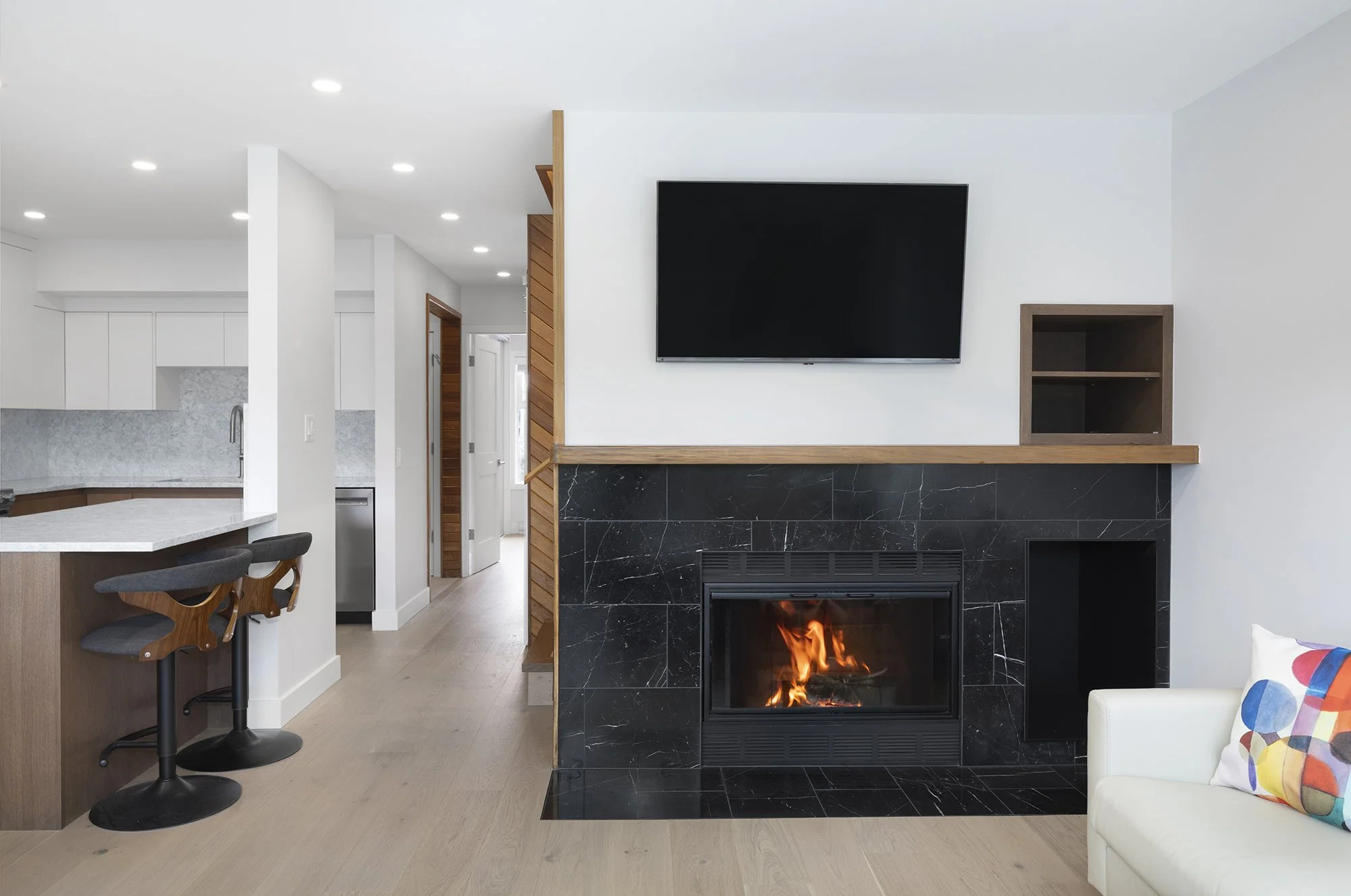 Modern living room with a fireplace, a flat-screen TV mounted on the wall, and a kitchen area in the background. The fireplace is black with gold veining and fire burning inside. The room has white walls, wood flooring, and contemporary furniture, in