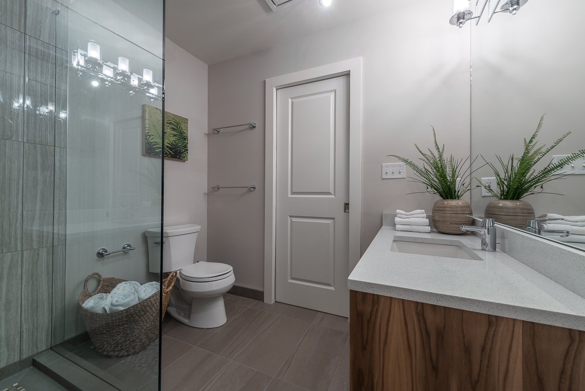 Modern bathroom with glass shower, white toilet, wooden vanity with white countertop, two potted plants, folded towels, and light gray tiled floor.