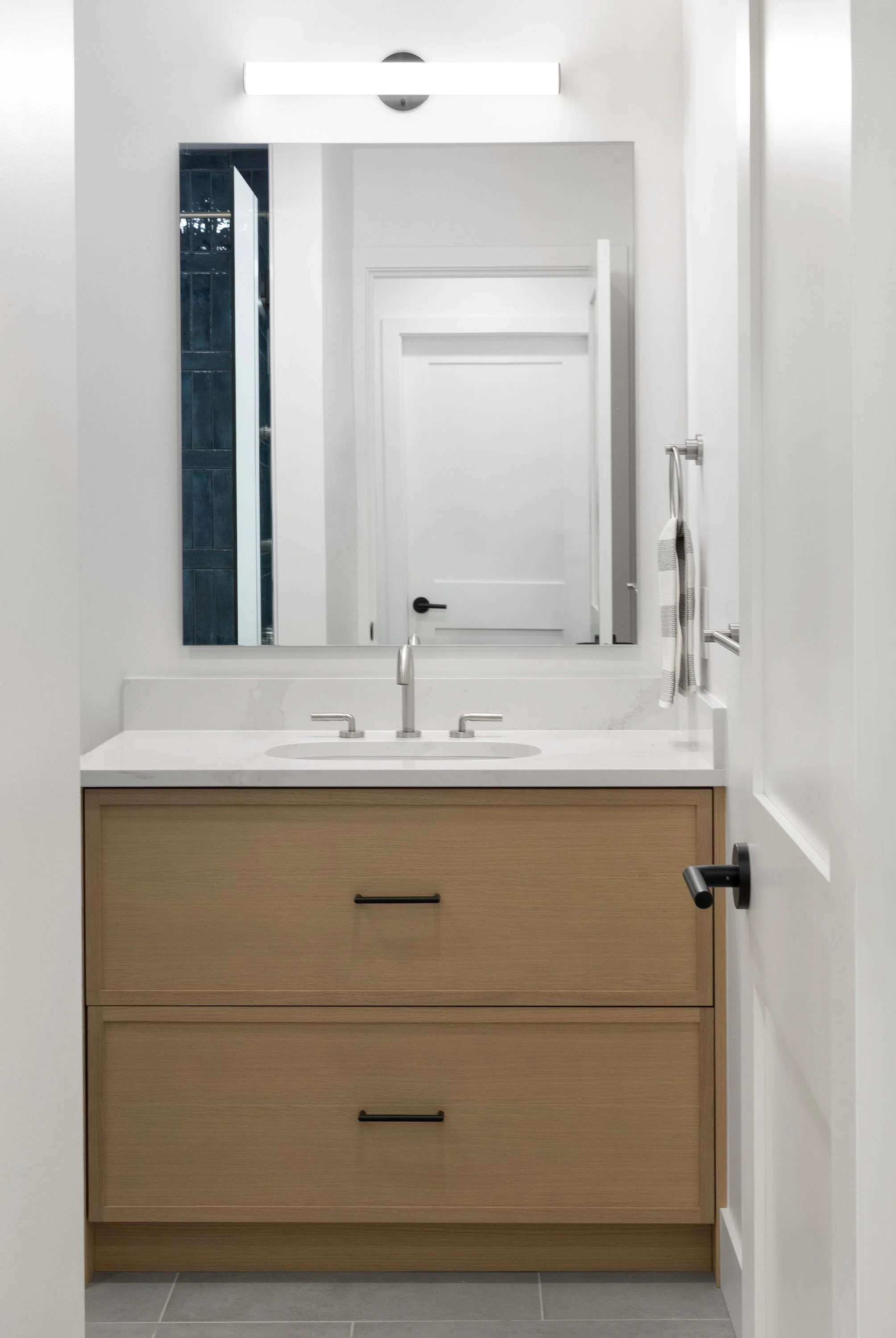 Modern bathroom mirror with a strip light above it, white walls, a white door, a beige wooden vanity with drawers, a white countertop, a sink, silver faucet, and a towel hanging on the wall.