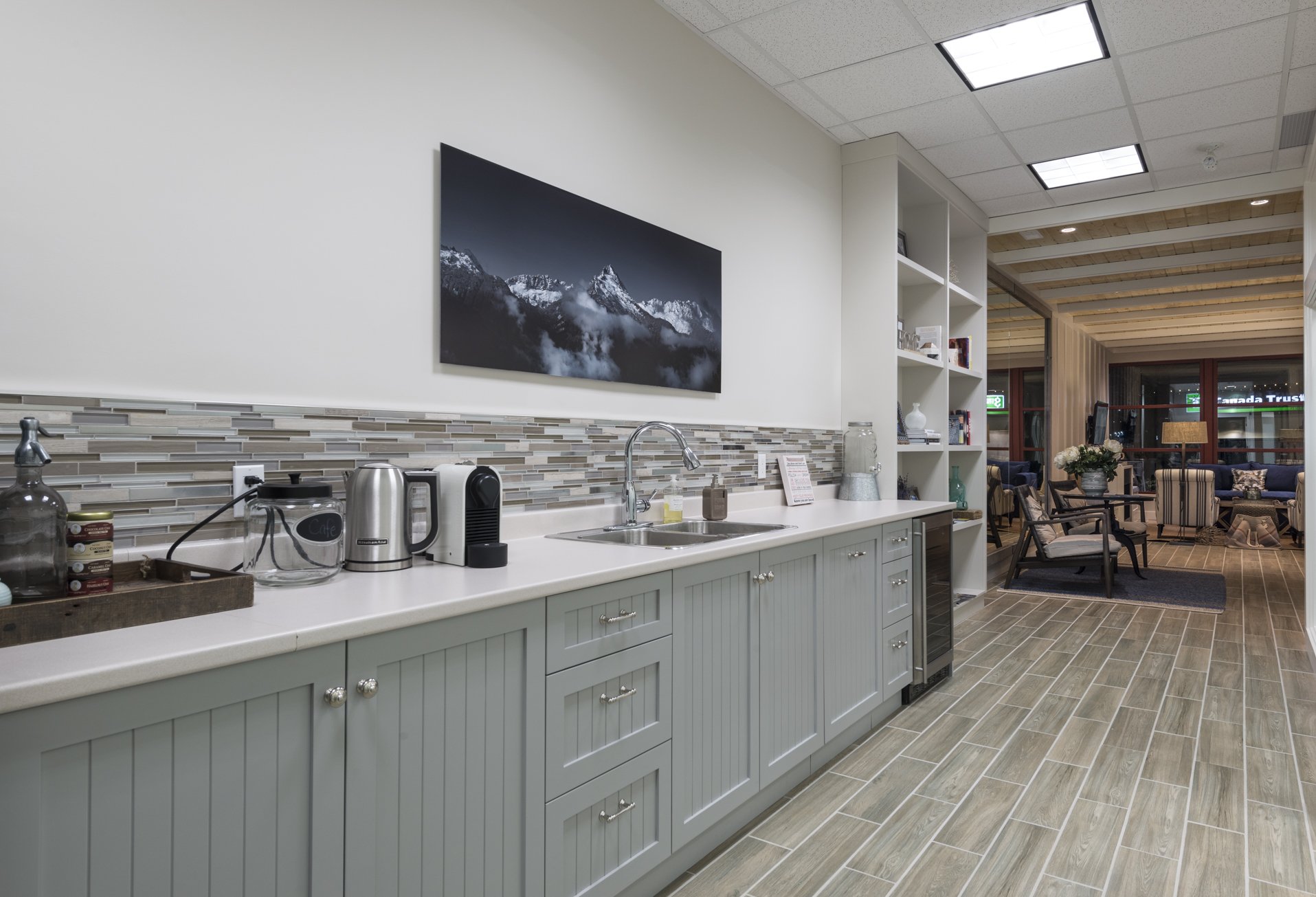 A kitchenette with teal cabinets, a white countertop, and a colorful tile backsplash. There are various kitchen appliances and items on the counter, a mountain landscape picture on the wall, and a seating area with chairs and a table in the backgroun