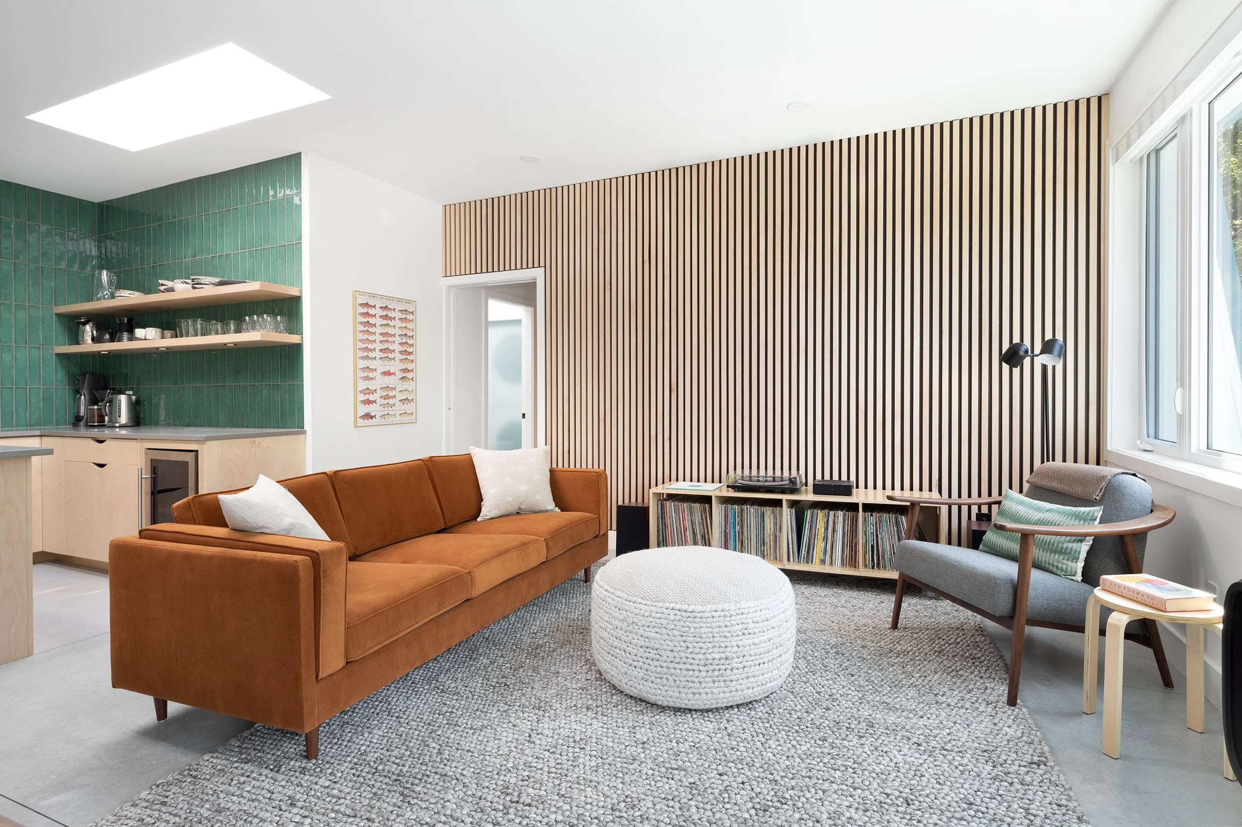 Living room with a brown sofa, a white pouf, a gray armchair, a wooden side table, a bookshelf with records, a wooden-textured accent wall, a window, and a ceiling light.