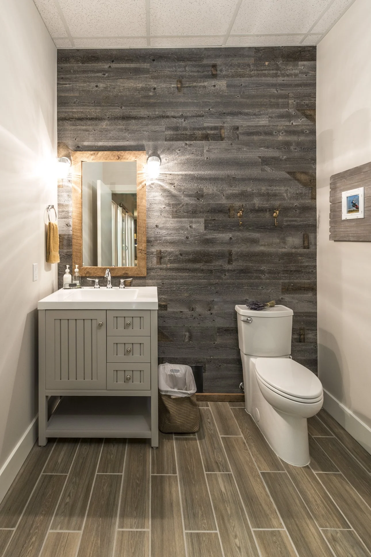 A small bathroom with wood-look tile flooring, a white toilet, a white vanity with a mirror, and a rustic wood accent wall with two small mounted hooks. There's a framed picture on the right wall, a towel on a ring on the left wall, and soap bottles 