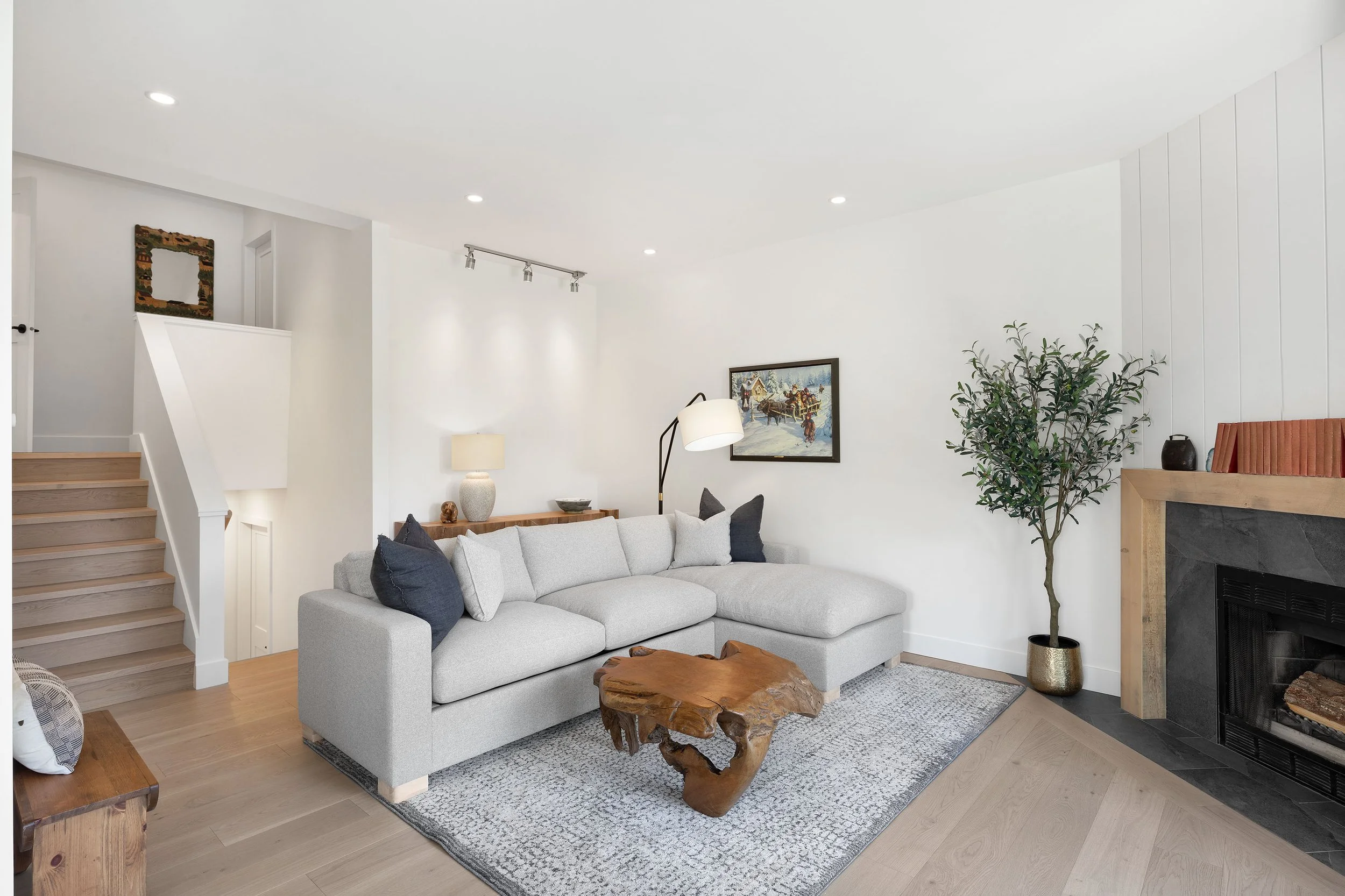 Living room with a light gray sectional sofa, wooden coffee table, fireplace, large green potted plant, wall art, lamps, and hardwood flooring.