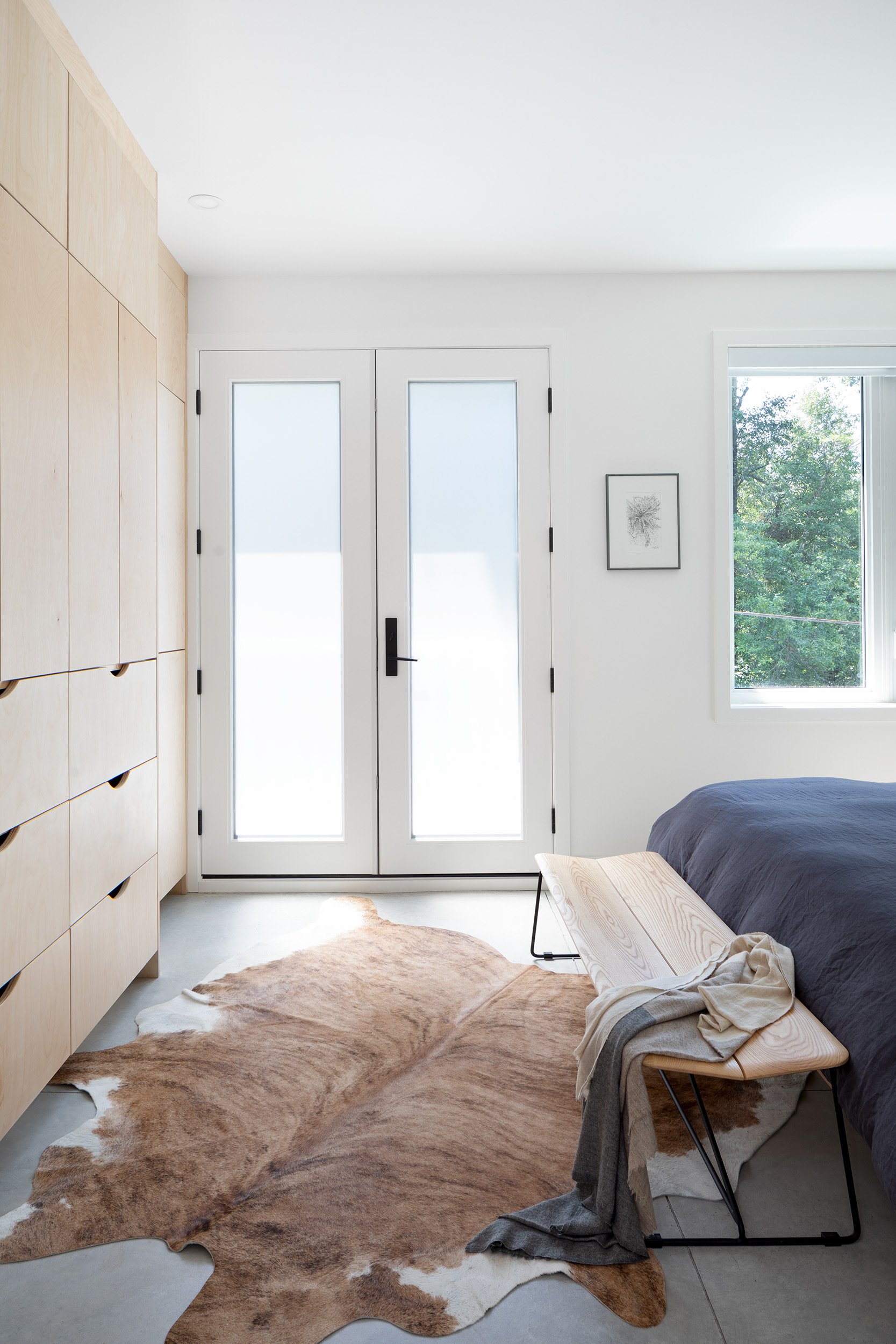 Modern bedroom with white walls, a large cowhide rug on the concrete floor, a wooden bench, a bed with dark gray bedding, a wooden dresser, framed art on the wall, and windows showing green trees outside.