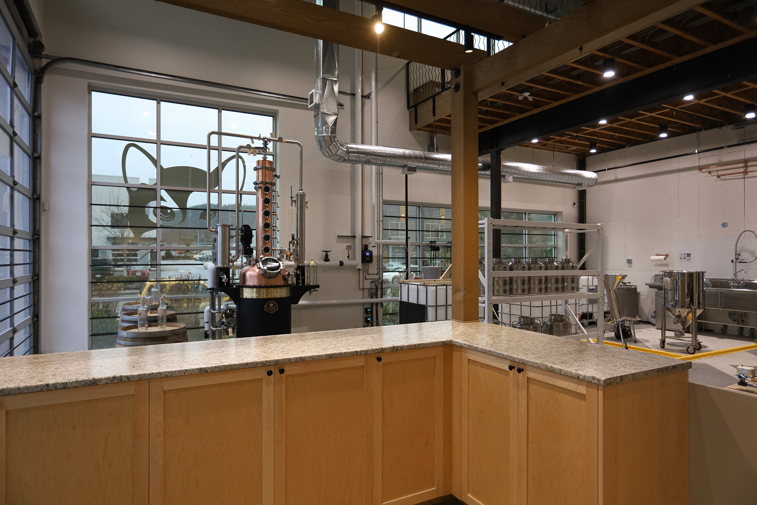 Interior of a brewery or distillery with brewing equipment, large windows, and a counter in the foreground.