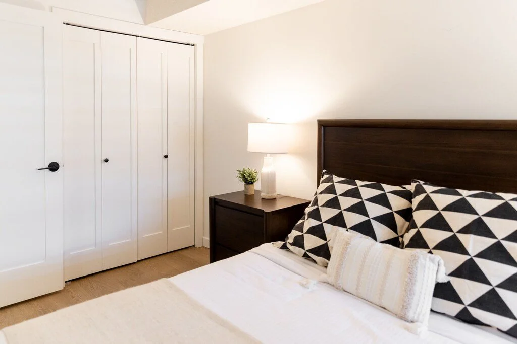 A bedroom with a bed, black and white geometric pillows, a bedside table with a lamp and a small plant, and a closet with white doors.
