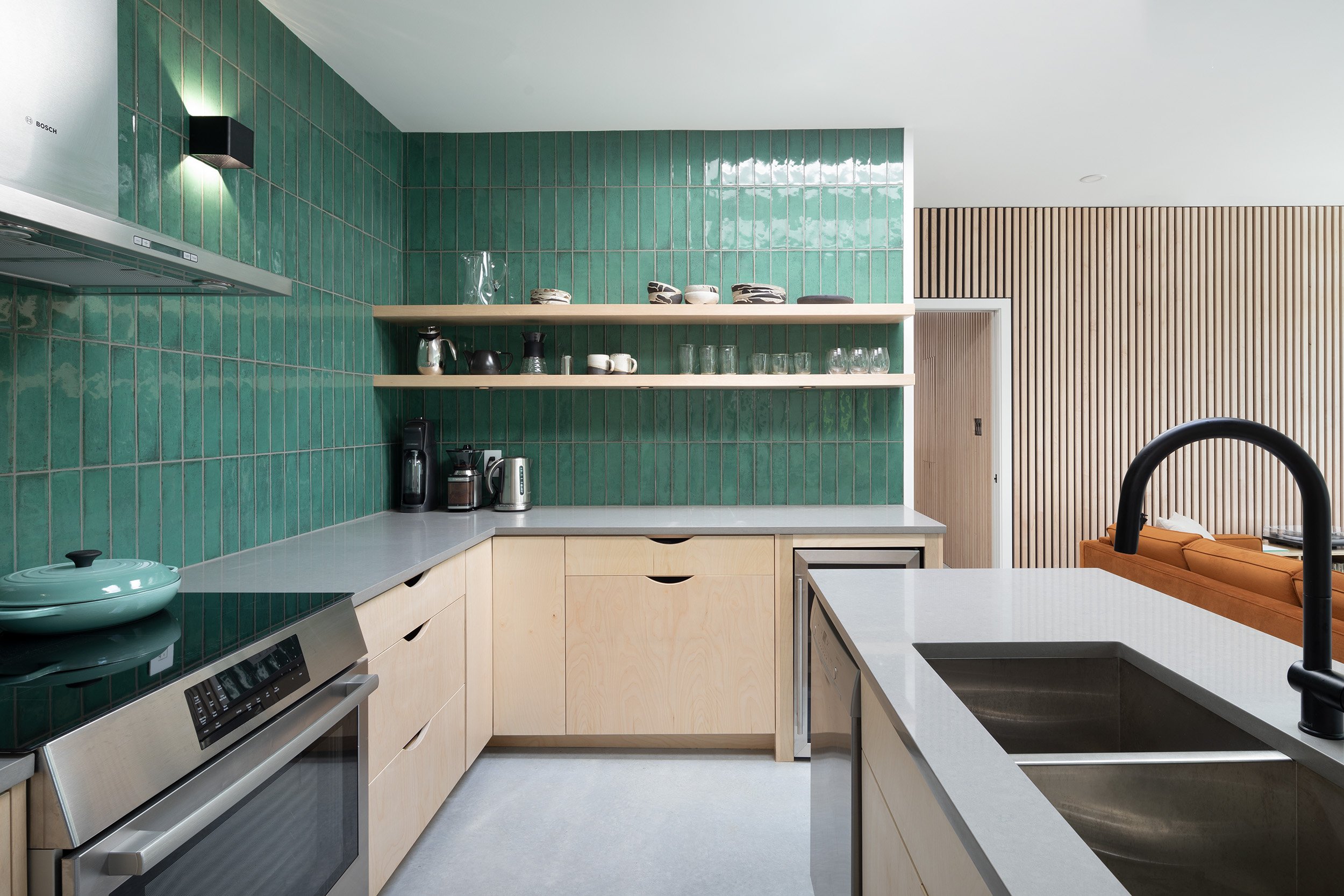 Modern kitchen with green tiled backsplash, light wood cabinets, and open shelves holding dishes, glasses, and kitchenware. includes a black faucet and a stovetop with a tea pot.