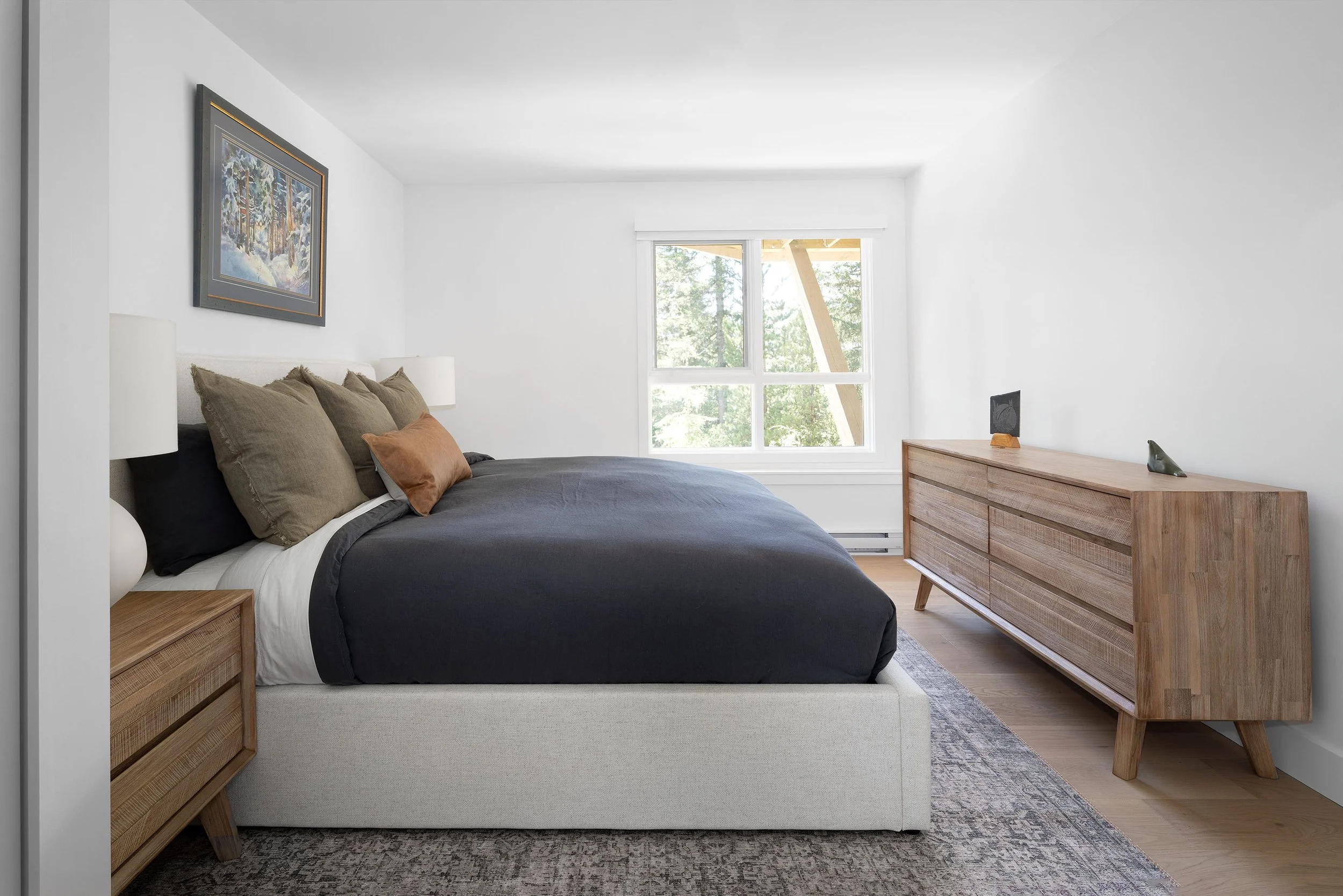 A bedroom with white walls, a large window showing trees outside, a bed with beige and dark gray bedding, and a wooden dresser with minimal decorations.