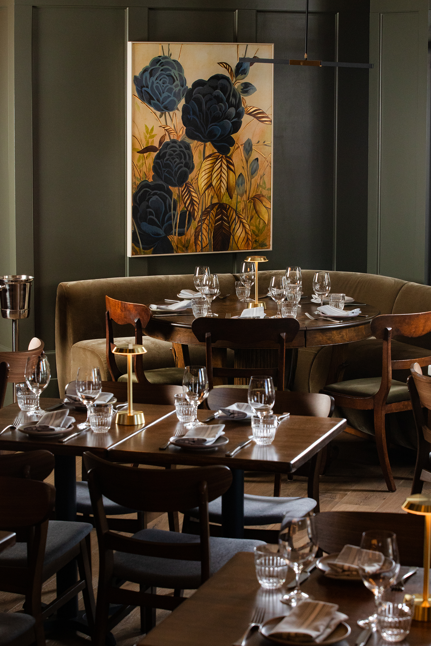 A cozy restaurant interior with a round wooden table set with glassware and napkins, surrounded by various wooden chairs and a curved booth, with a large floral painting with dark blues and earthy tones on a dark wall.