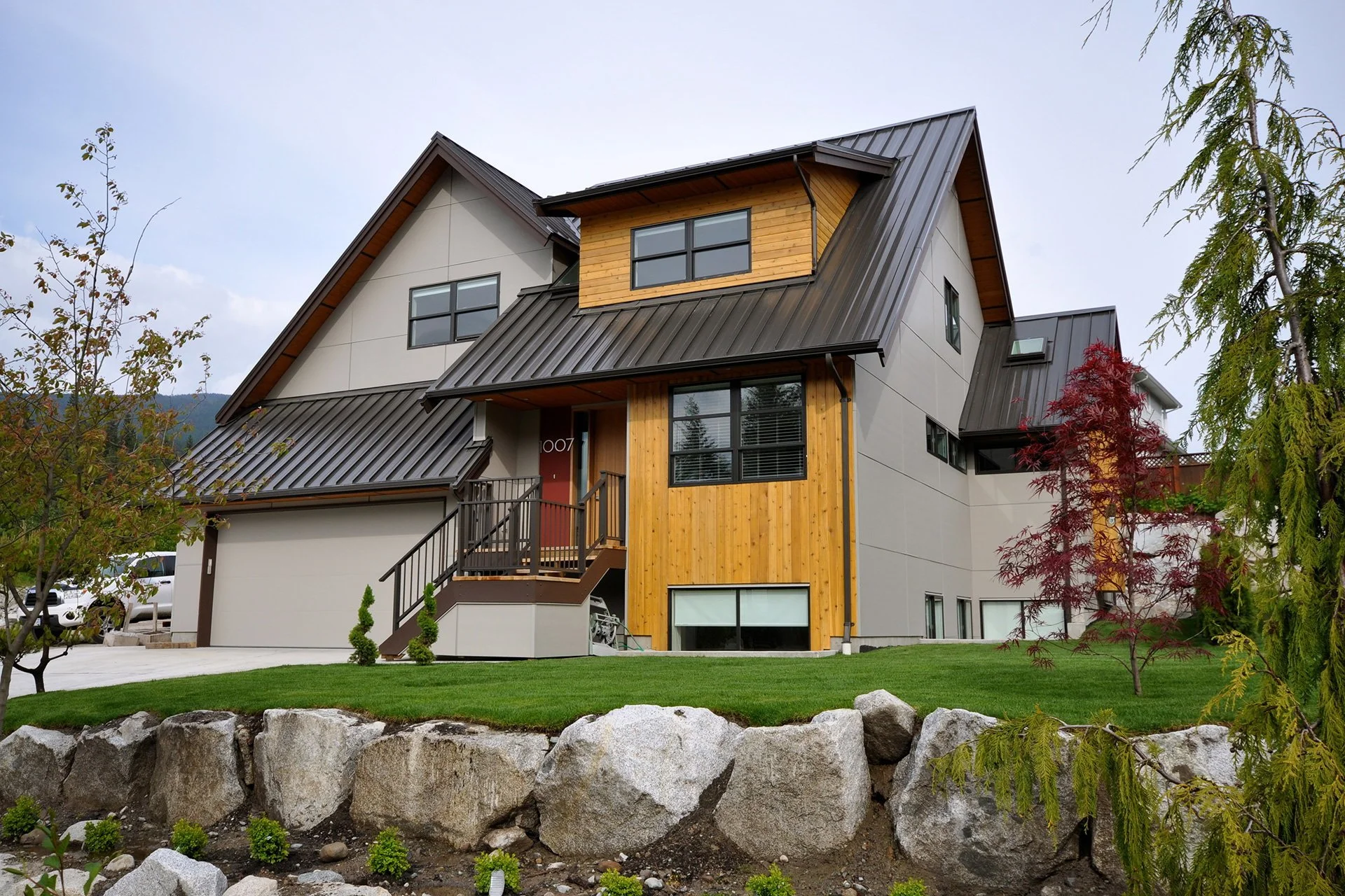 Modern multi-story house with a metal roof, wooden siding, and large windows, surrounded by landscaped yard and trees.
