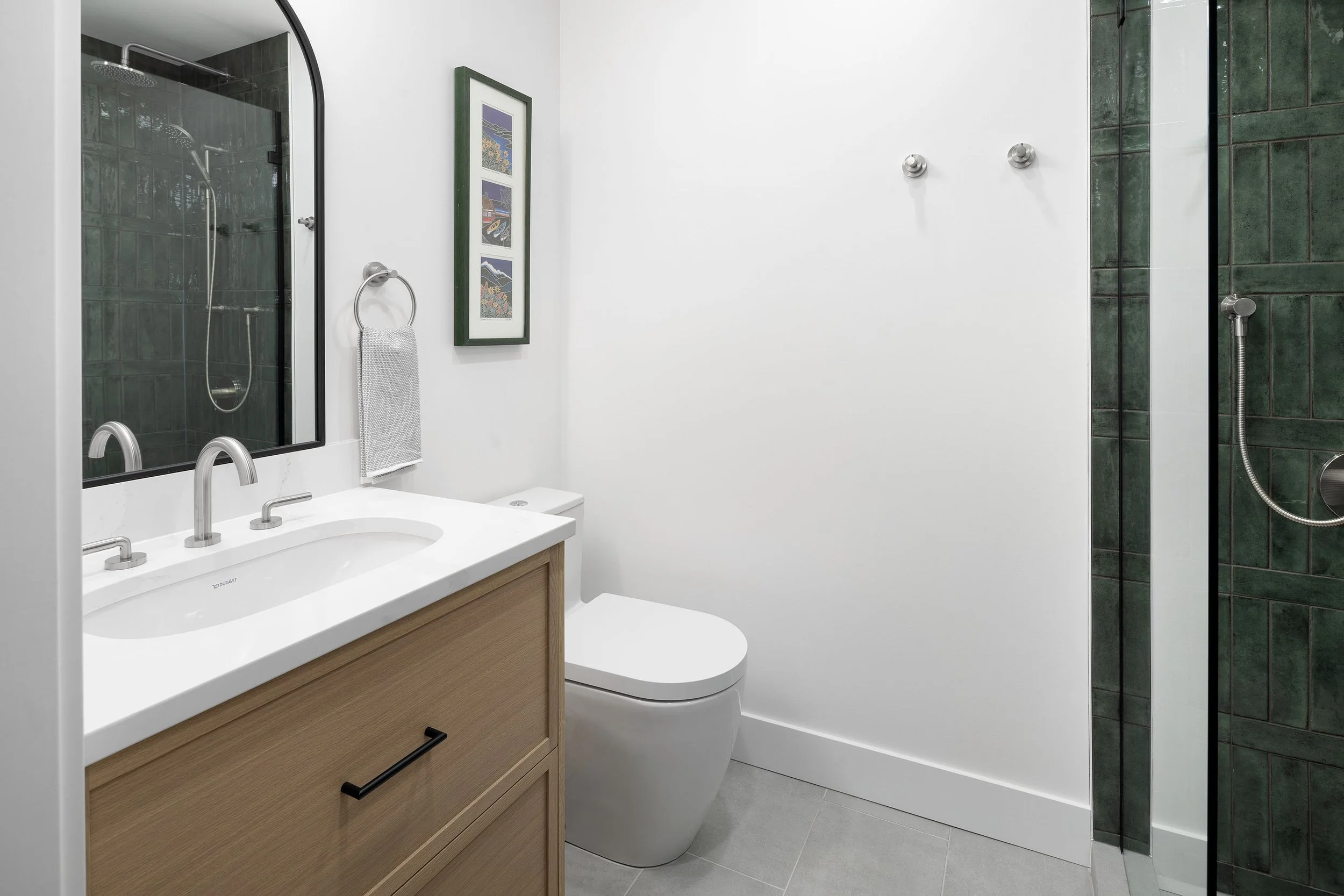 Modern bathroom with white walls, green tiled shower area, wooden vanity with cabinet, white toilet, and framed artwork on the wall.
