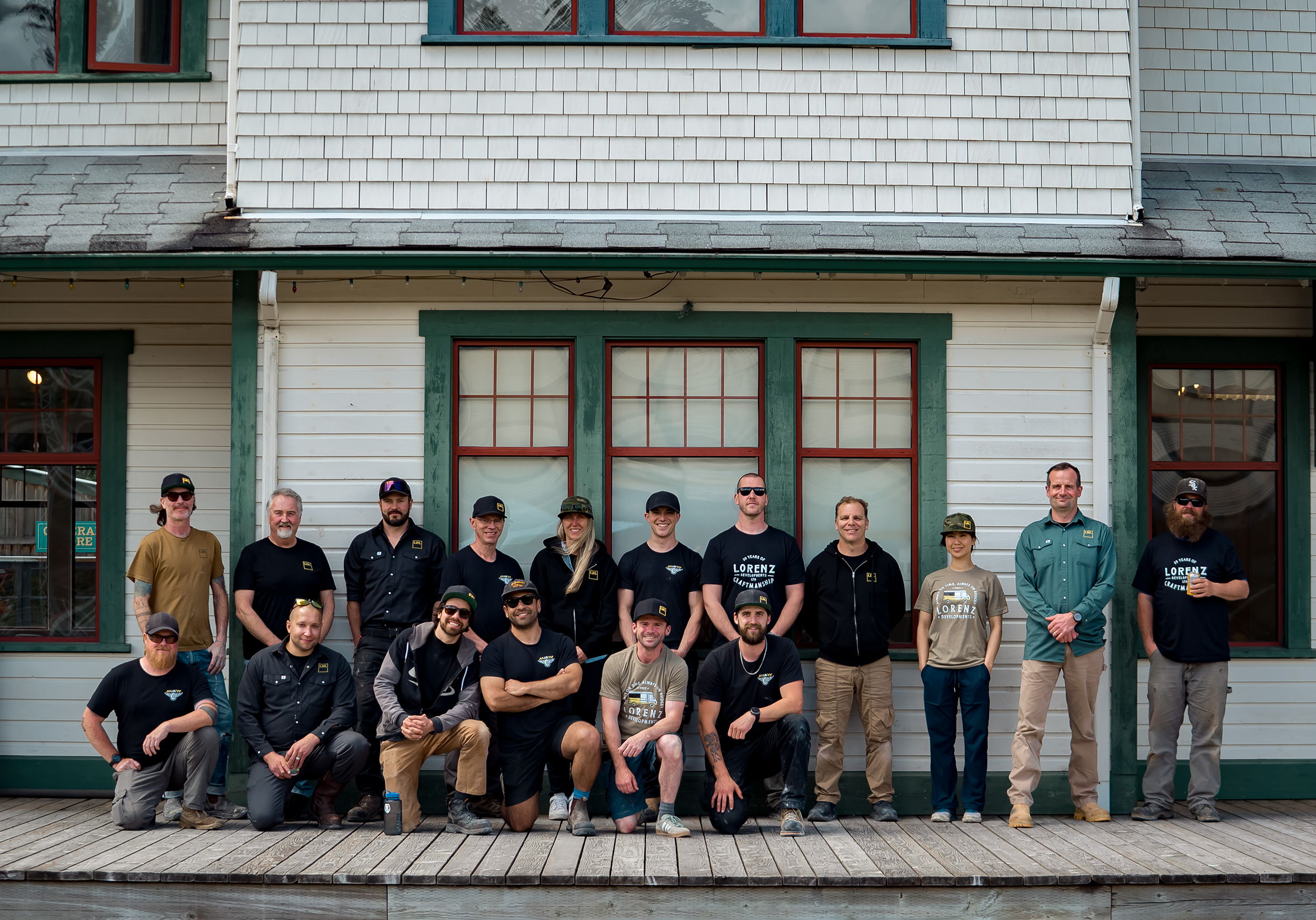 Group of people standing and kneeling outside a building with white siding and red window frames, some wearing sunglasses and casual clothing, smiling for a photo.