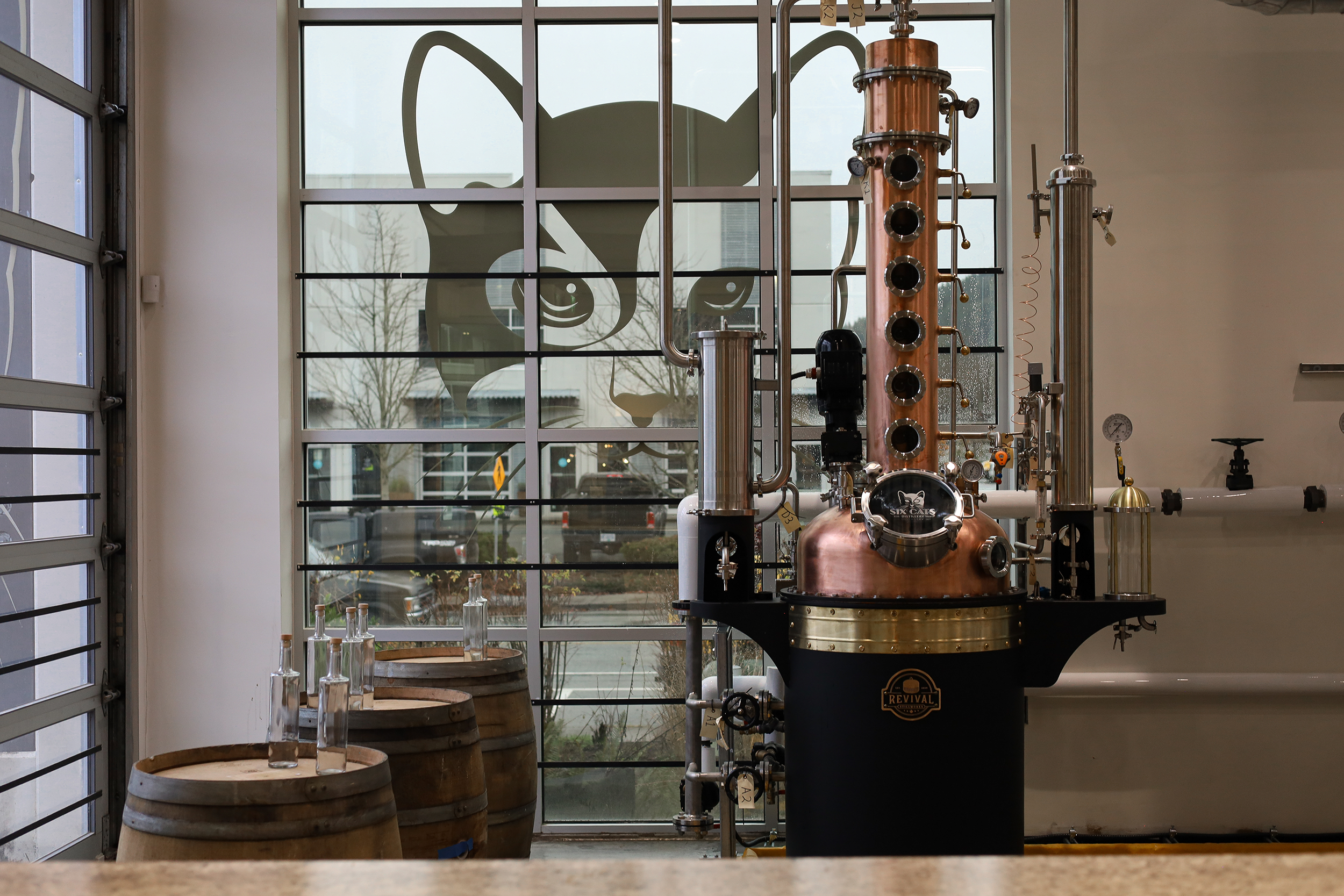 Inside a distillery with copper stills and glass bottles on wooden barrels, seen through large windows with a building and trees outside.