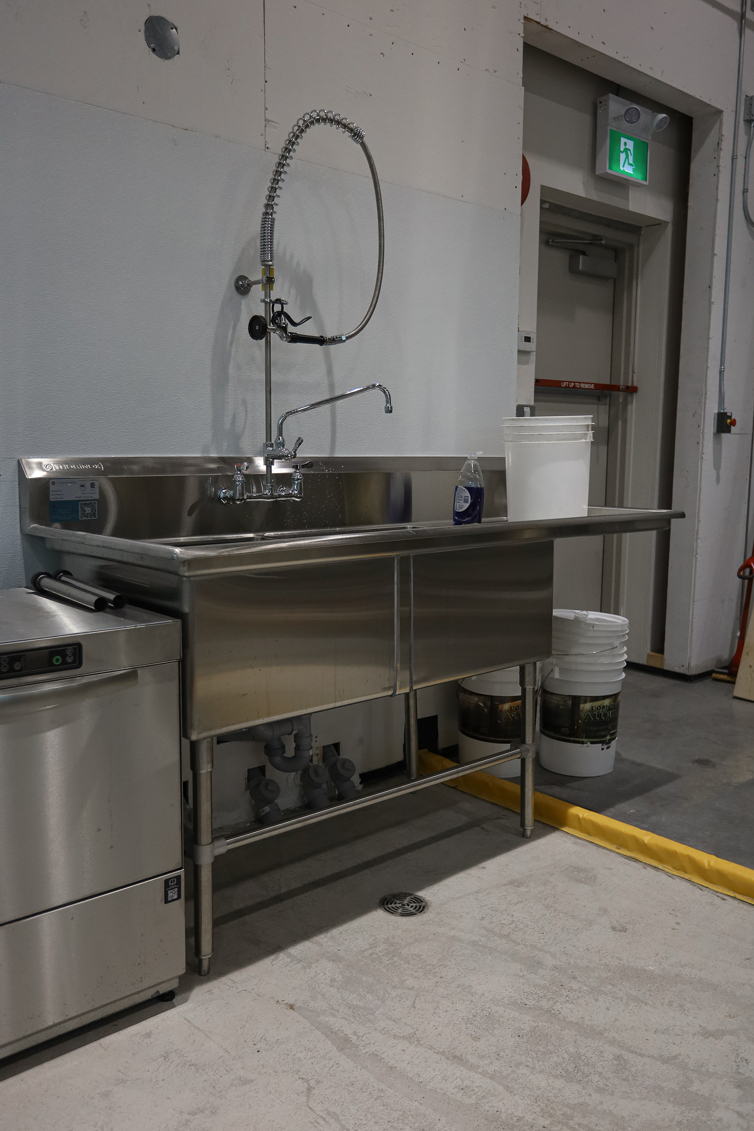A stainless steel utility sink with a flexible sprayer hose, a bottle of cleaning solution, and white buckets beneath it. In the background, there is an emergency exit sign above a door with a red bar indicating 'LIFT UP TO REMOVE' and a yellow cauti