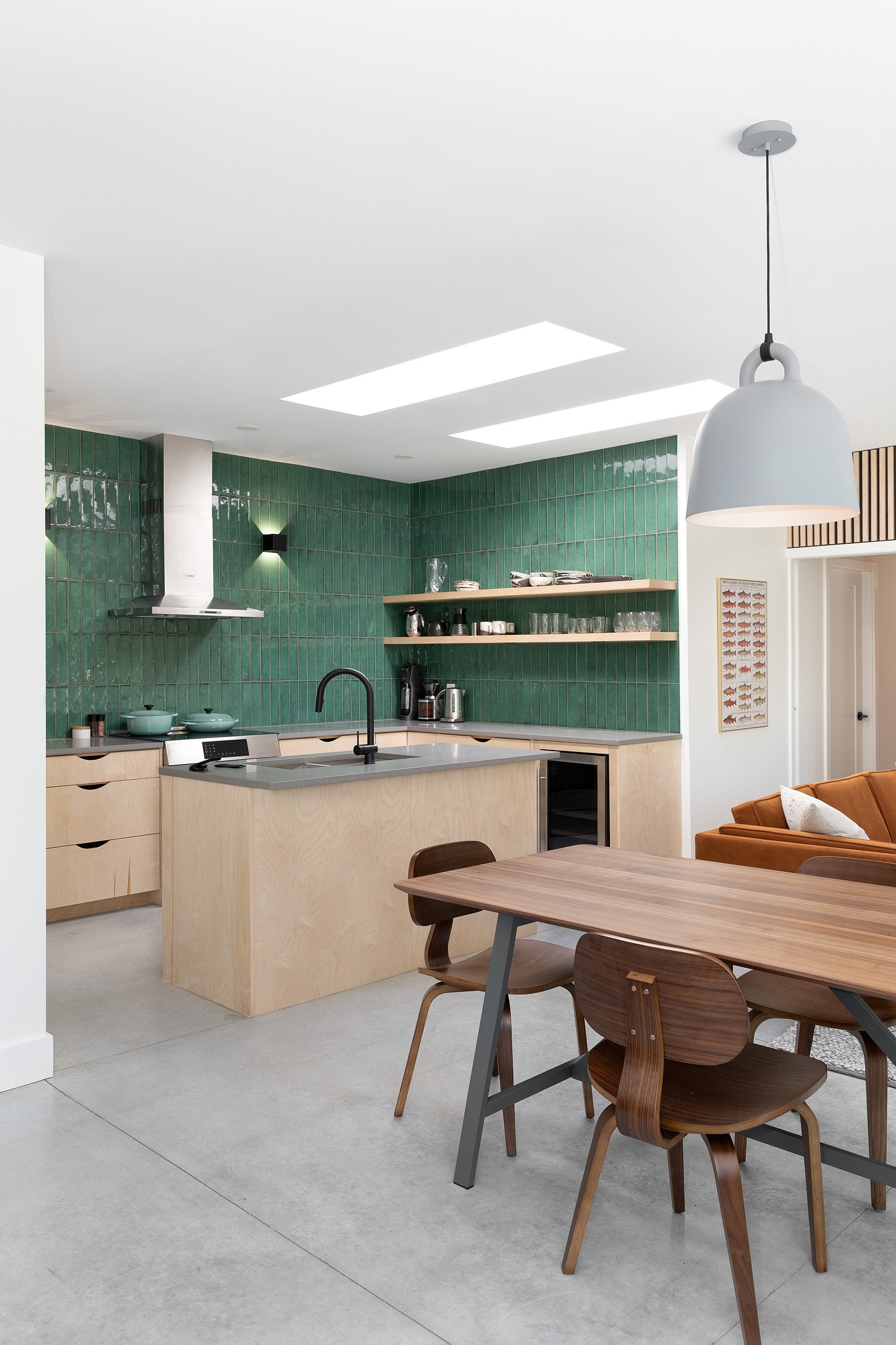Modern kitchen with green tiled wall, wooden cabinets, open shelves, stainless steel appliances, and wooden dining table with chairs, illuminated by natural light from skylights and a pendant lamp.