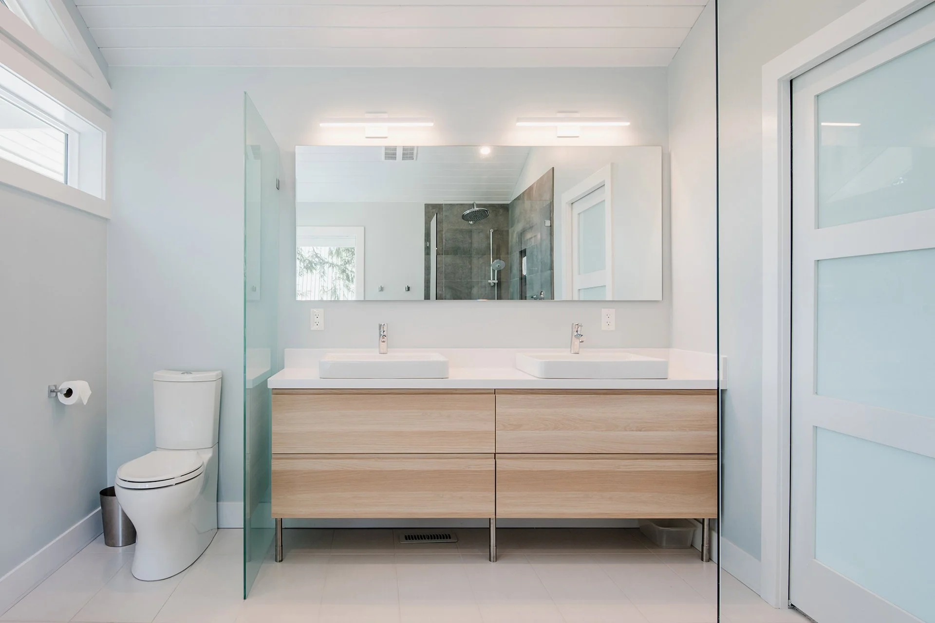 Modern bathroom with double sinks, large mirror, white walls, and a walk-in shower with glass door and gray tiles.