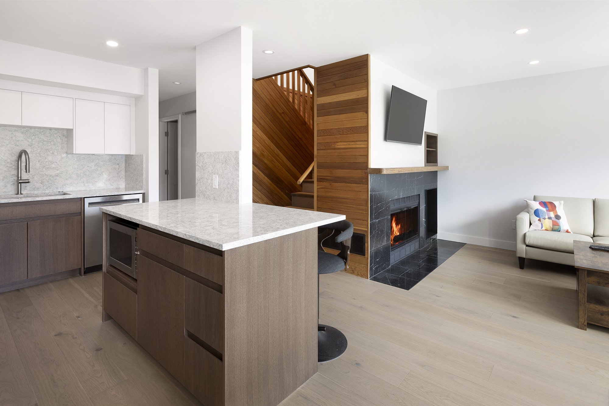 Open-concept kitchen and living room with white walls, wooden flooring, a kitchen island with a light gray countertop, a fireplace with a black marble surround and burning fire, a flat-screen TV on the wall, a white sofa with colorful pillows, and a 