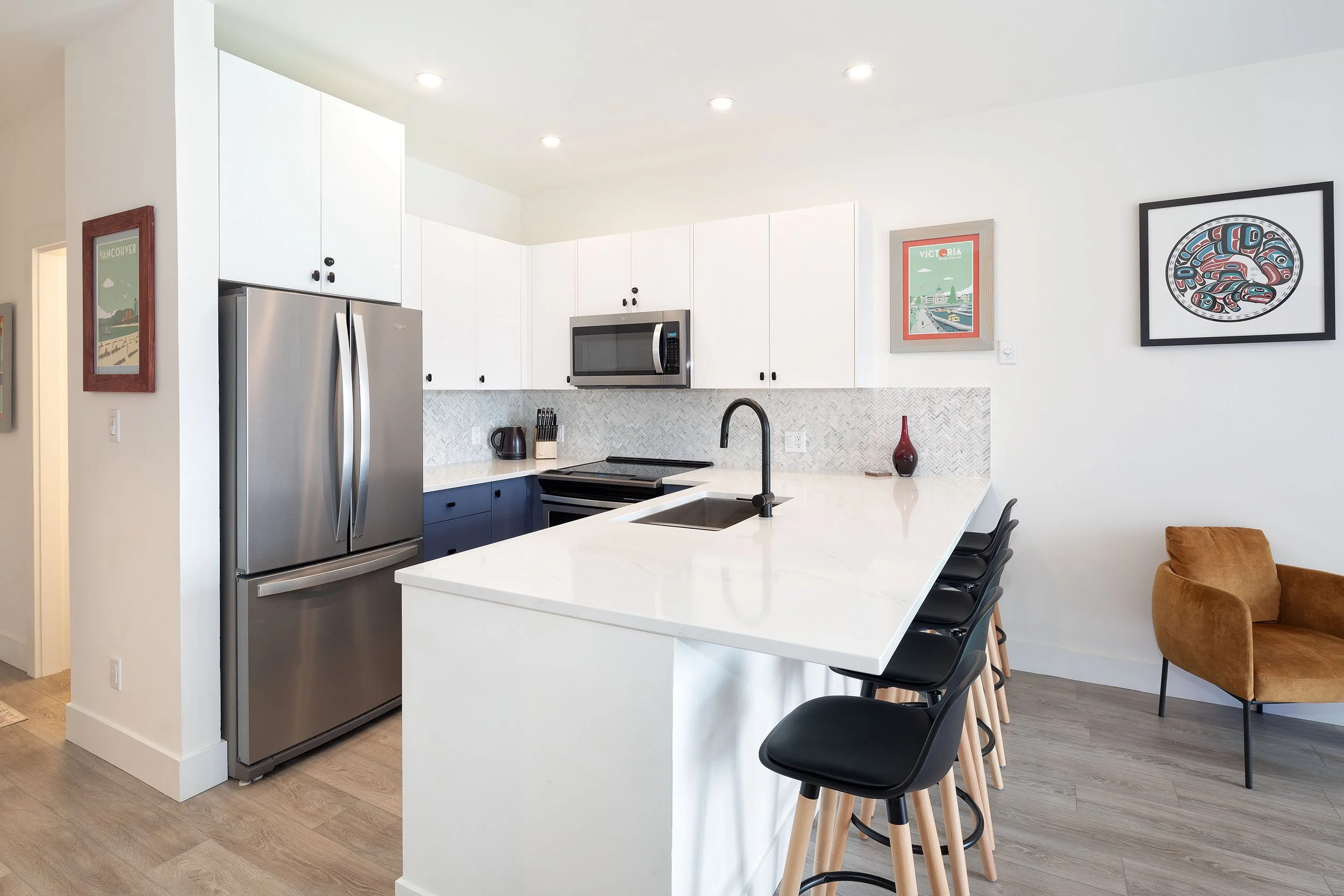 Modern kitchen with white cabinets, stainless steel refrigerator, black chairs at a white island, artwork on the wall, and light wood flooring.