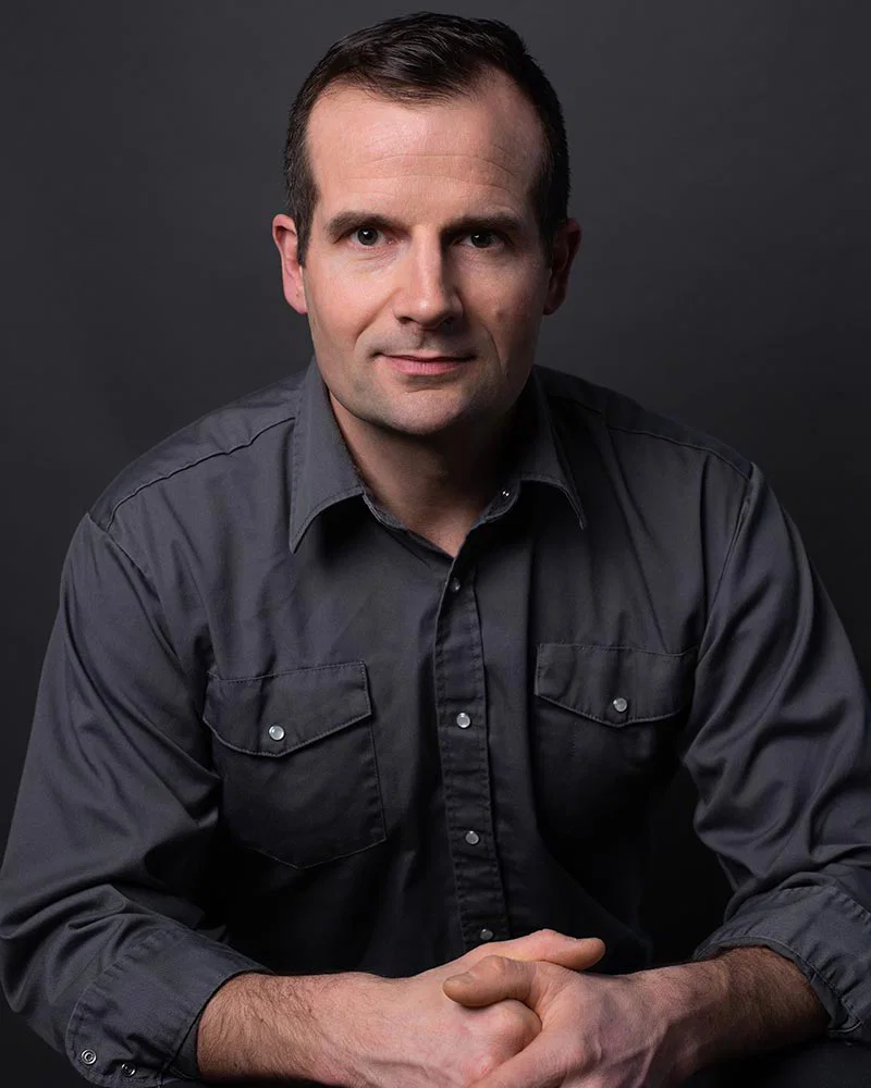 Portrait of a man with short dark hair, wearing a dark gray button-up shirt, sitting against a dark background.
